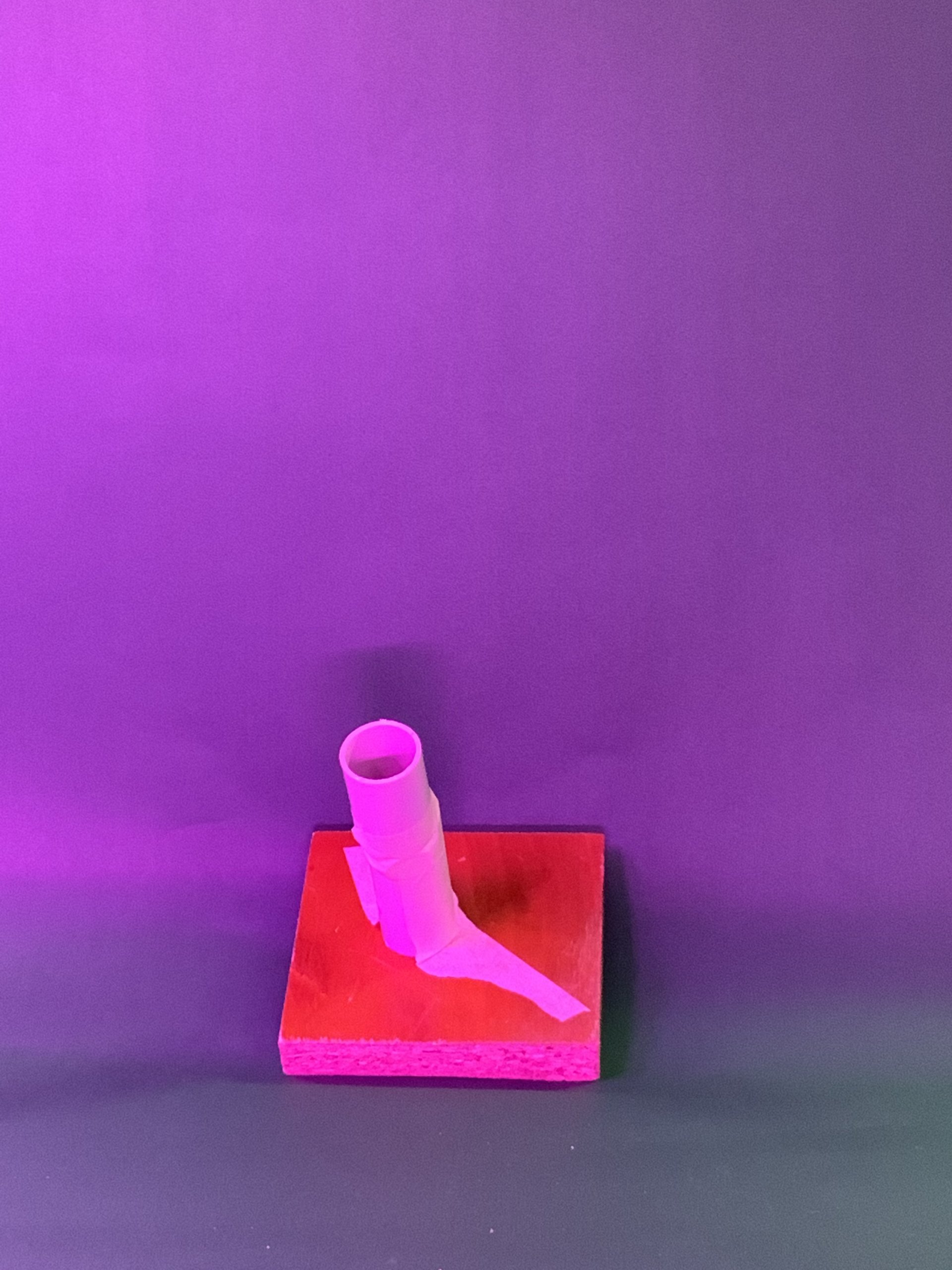 Un objet cylindrique avec une pièce allongée est monté sur une base rectangulaire rouge, sur un fond violet avec un éclairage doux.