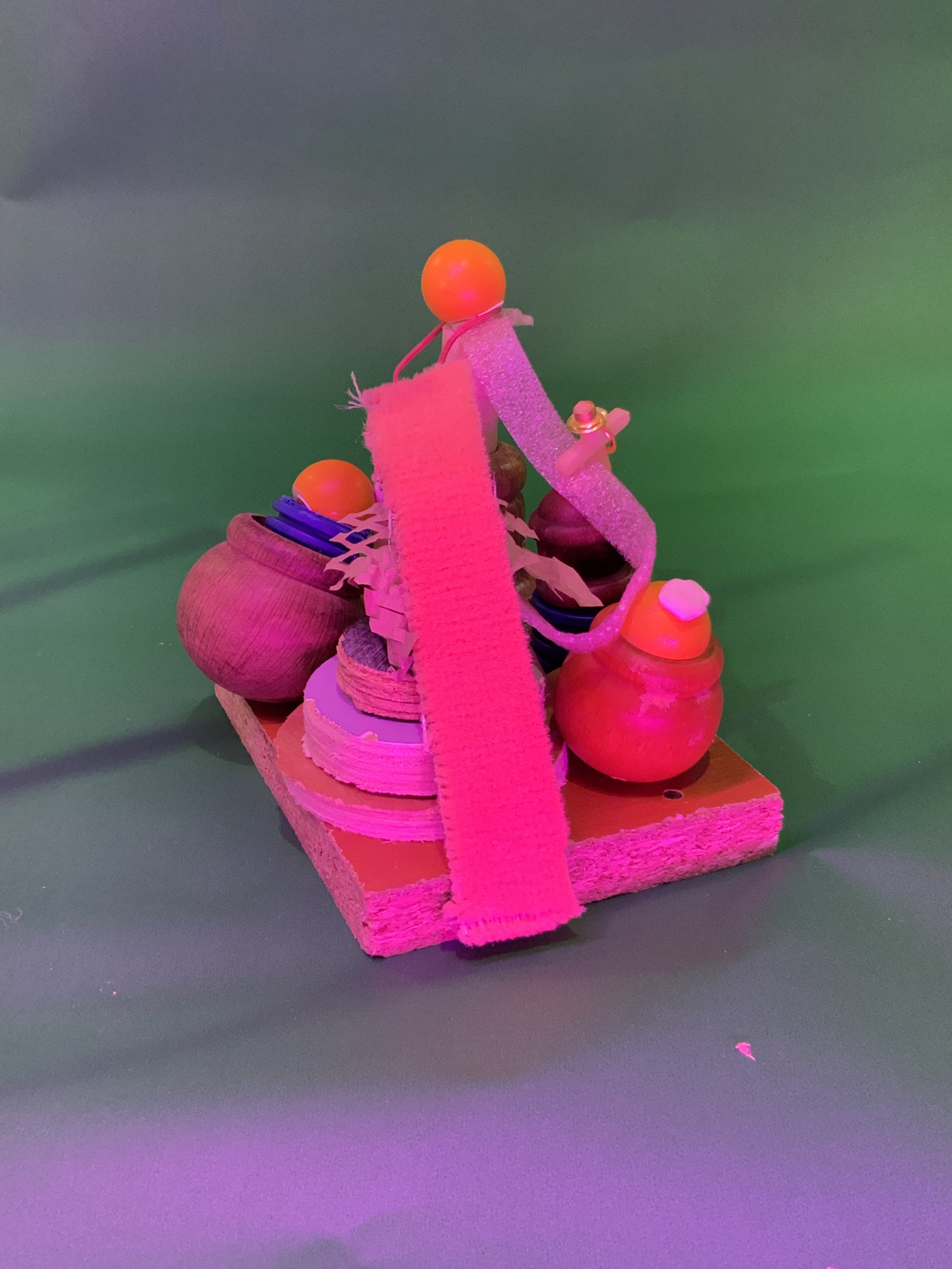 Une petite sculpture abstraite composée de formes en bois, de mousse, de boules en plastique et d'une bande de tissu rose, le tout assemblé sur une base rectangulaire en bois sur un fond vert avec un éclairage violet.