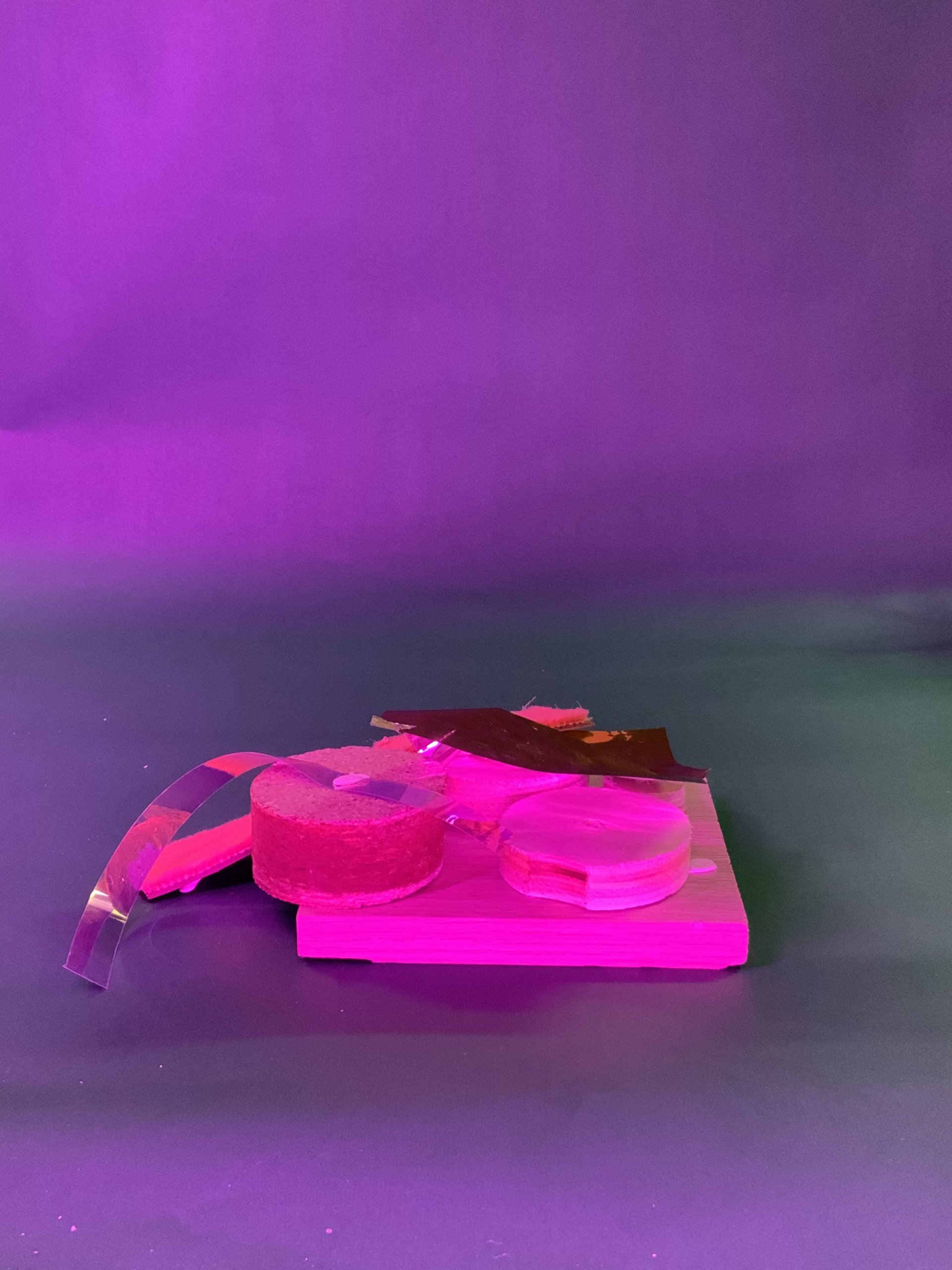 Un panneau en bois présente divers articles, dont deux rouleaux de ruban adhésif, une bande de plastique transparente et un morceau de matériau rouge réfléchissant, le tout sous un éclairage rose et violet sur un fond uni.