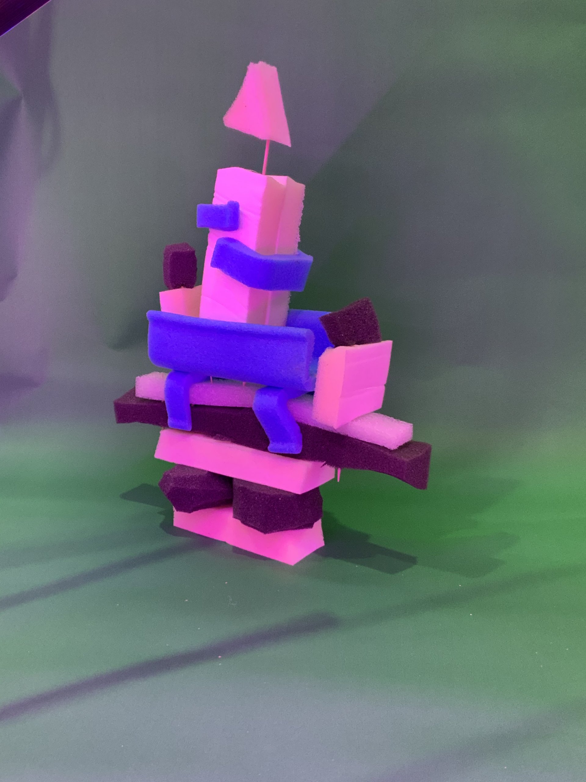 Une pile d'éponges colorées, roses, bleues et noires, est disposée selon une structure géométrique abstraite sur fond vert. Une petite éponge rose triangulaire trône au sommet.