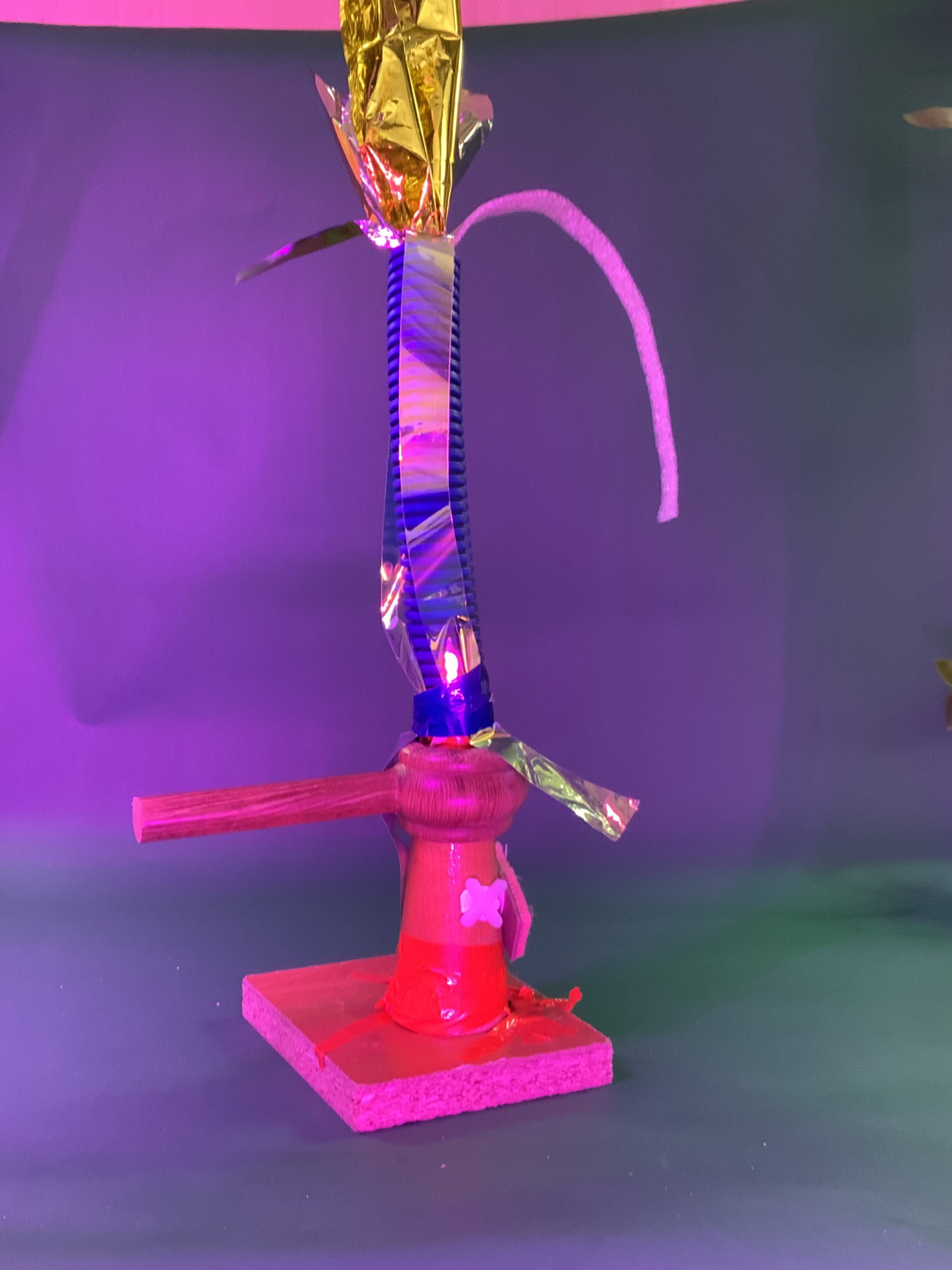 Une sculpture réalisée à partir de divers matériaux, dont une bobine en bois, une plate-forme rouge, un morceau de bois, du plastique, une feuille d'or métallique et un cure-pipe blanc, est présentée sur un fond violet et vert.