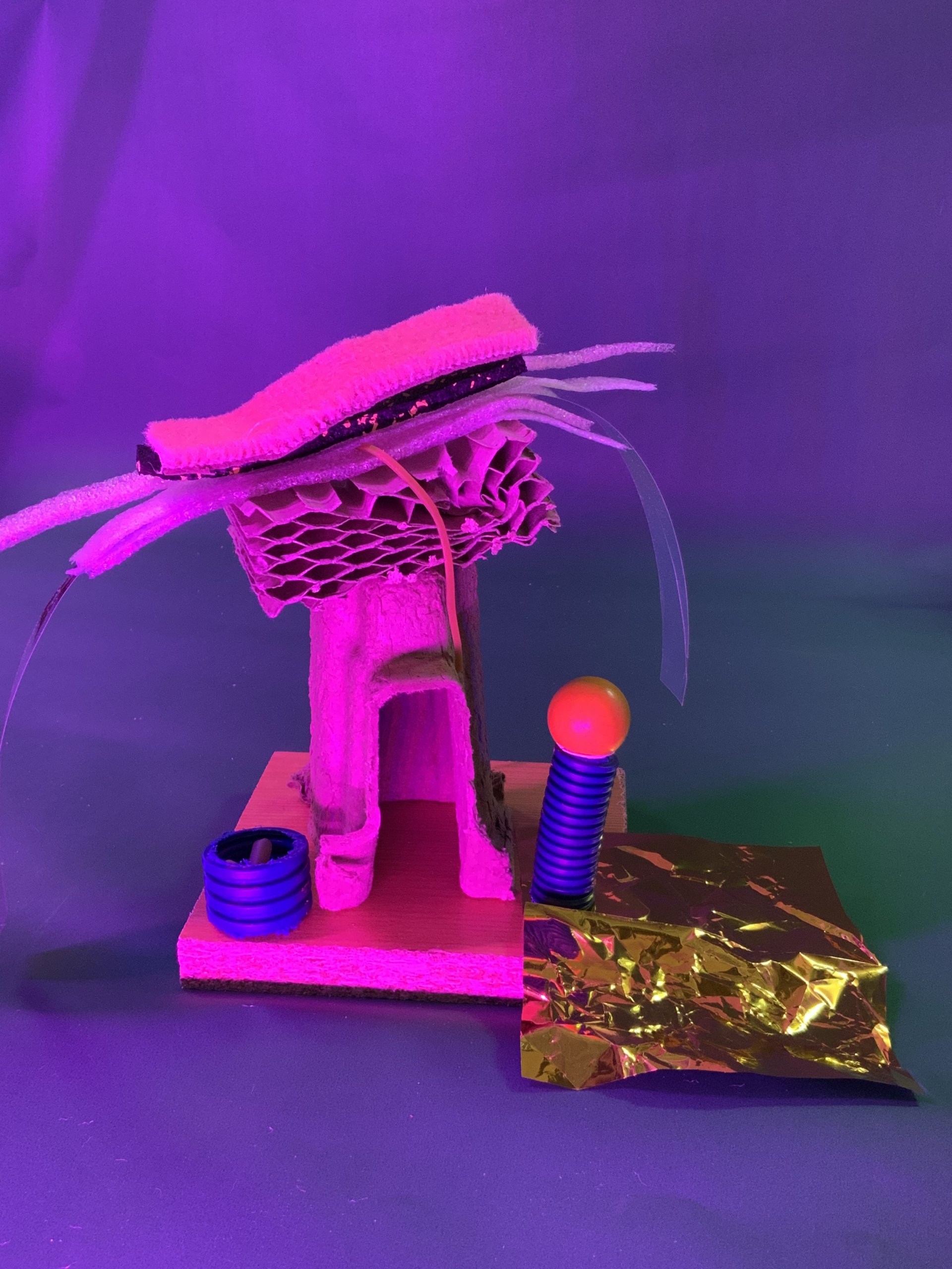 Une petite sculpture abstraite réalisée à partir de carton, de papier nid d'abeille, de feutre, de tubes en plastique, d'une bobine bleue, d'un ressort avec une boule orange sur le dessus et d'une feuille d'or, le tout sur une base en bois avec un éclairage violet.