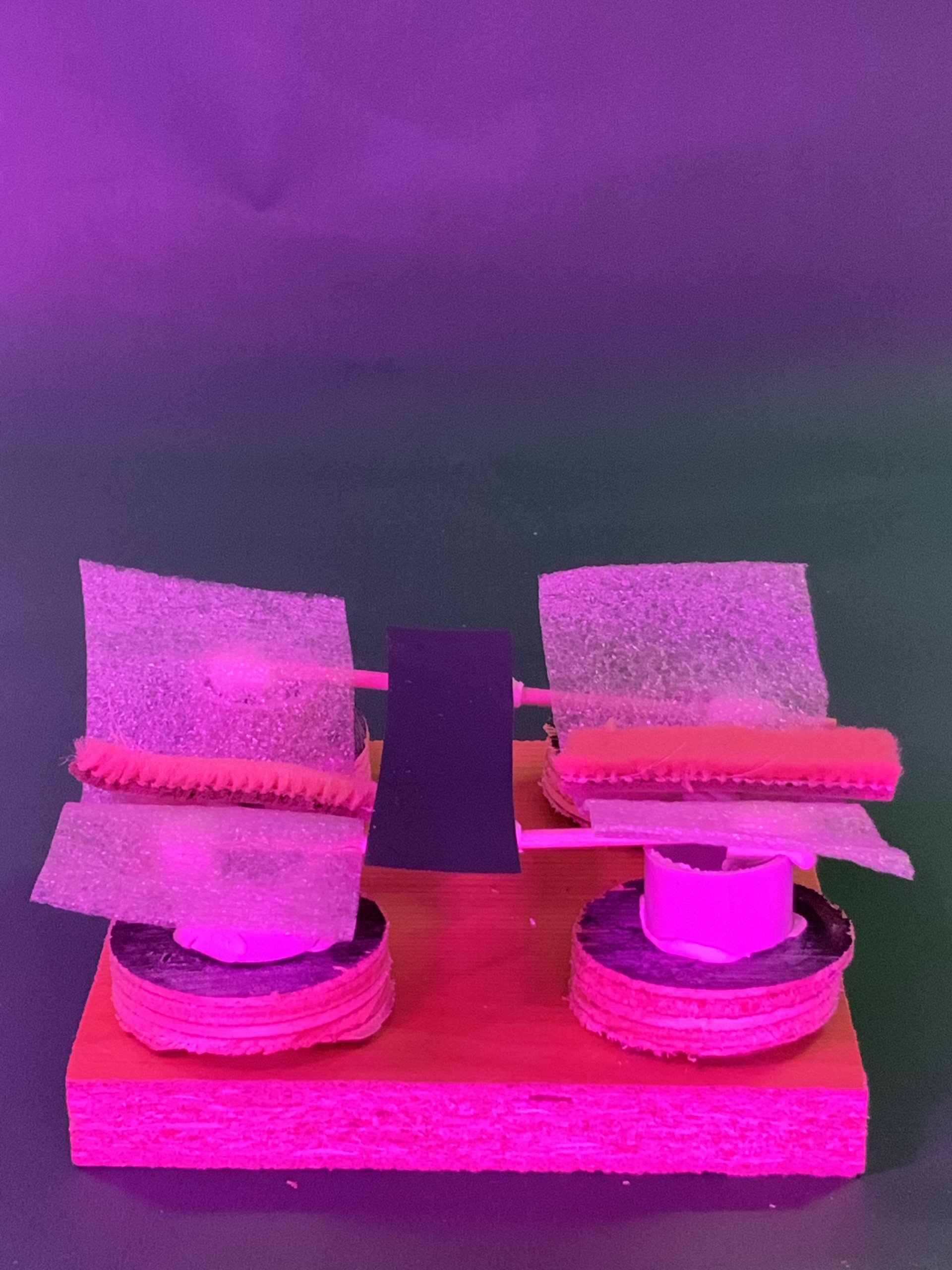 Une petite maquette artisanale repose sur un socle en bois composé de deux piliers composés de disques superposés, supportant des pièces inclinées en matériau translucide fixées par des tiges. La scène est éclairée par un fond dégradé rose et vert.