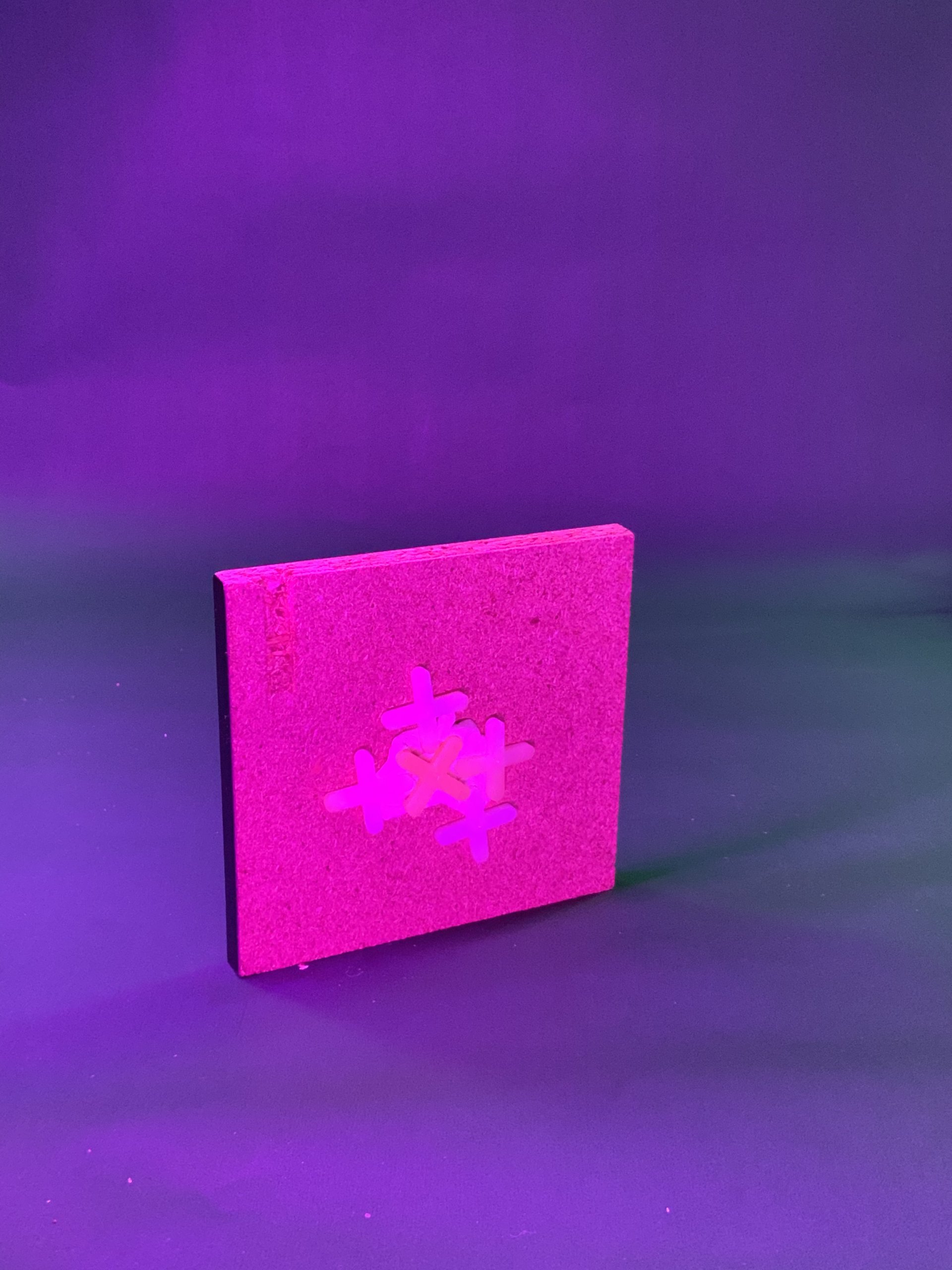 Un carreau rose texturé et carré se dresse sur un fond violet. Au centre, un symbole géométrique ressemblant à des croix ou des signes plus entrelacés. L'éclairage est vif et majoritairement violet.