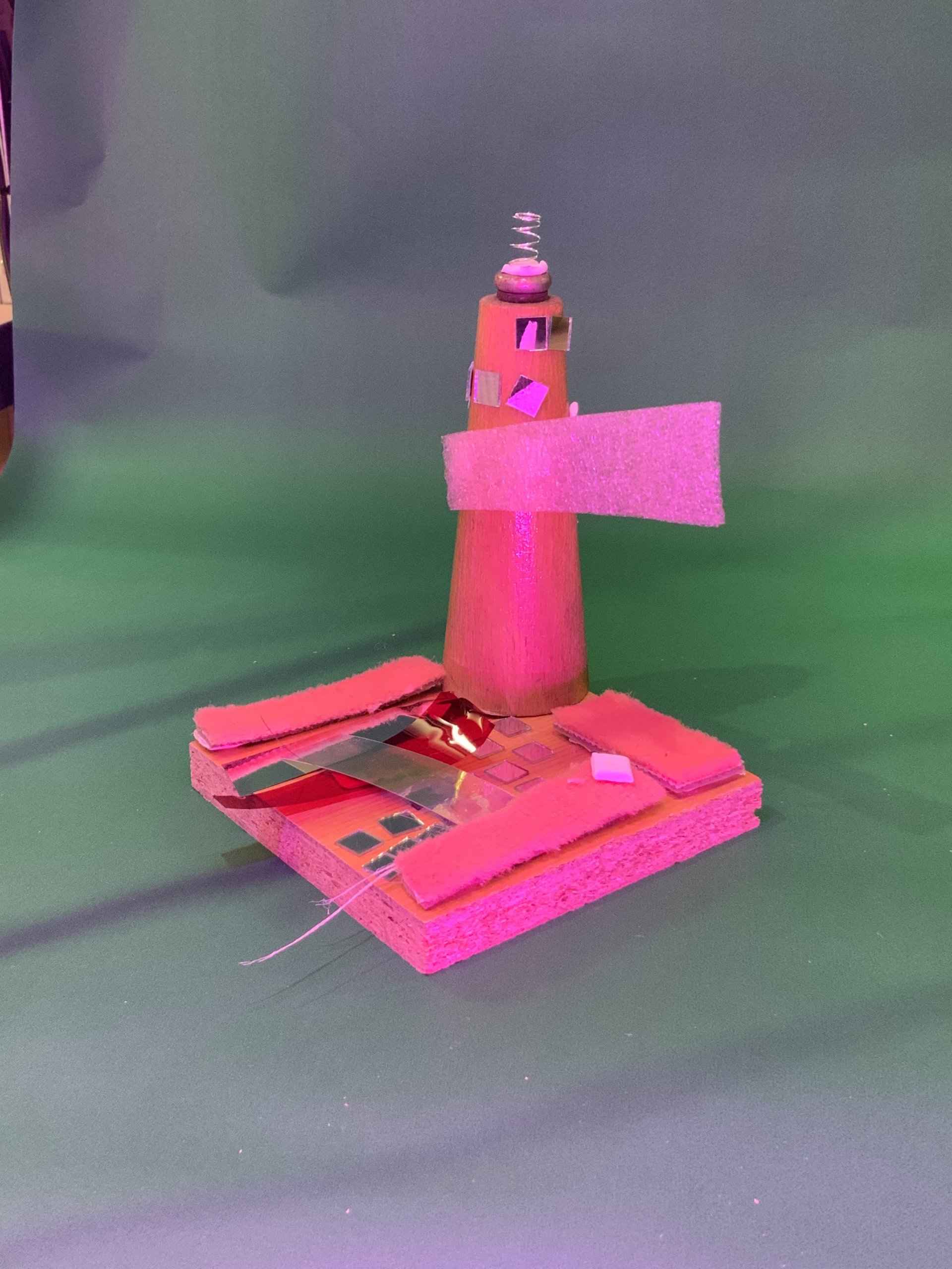 Modèle fait main représentant un phare en mousse rose avec un dessus en métal en spirale, monté sur une base rectangulaire décorée de petits miroirs et de pièces en mousse rose, sur un fond vert.