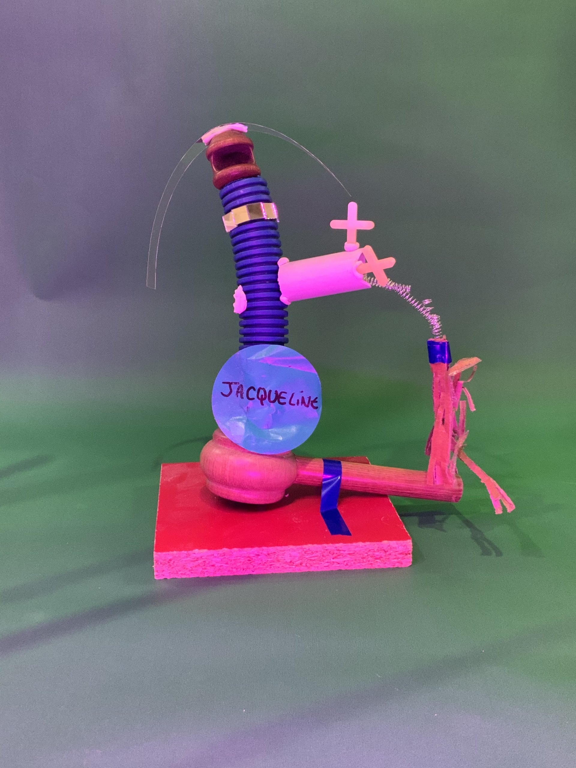 Une sculpture mixte composée d'un tube bleu, de pièces en plastique roses, d'une base en bois, d'un ruban et d'une boule bleue étiquetée Jacqueline, présentée sur un fond dégradé vert et violet.