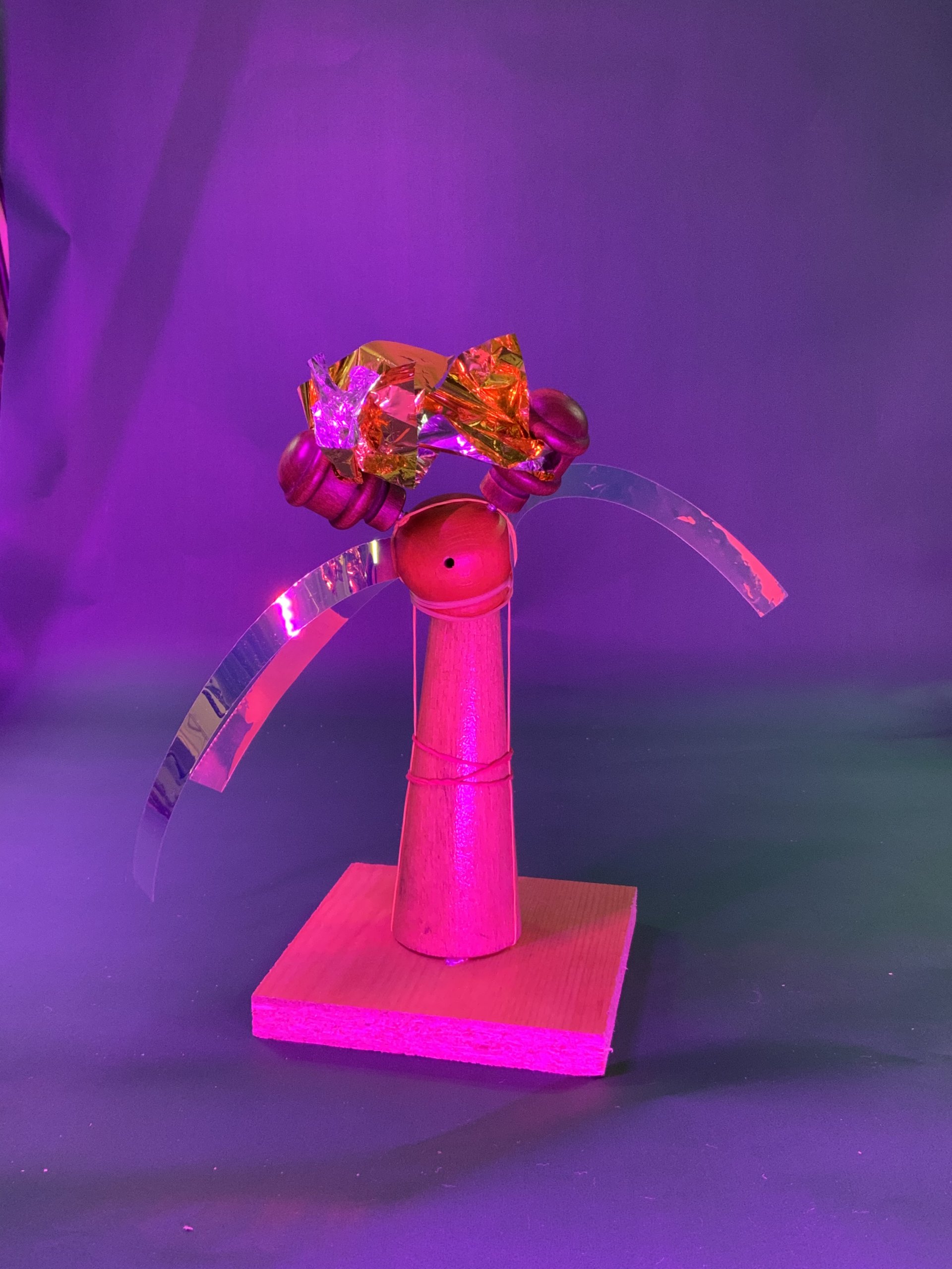 Une figurine en bois à la tête ronde et au corps conique repose sur un socle carré. La tête est surmontée de bonbons emballés et de bandes métalliques incurvées. Le fond est violet avec un éclairage rose et violet.