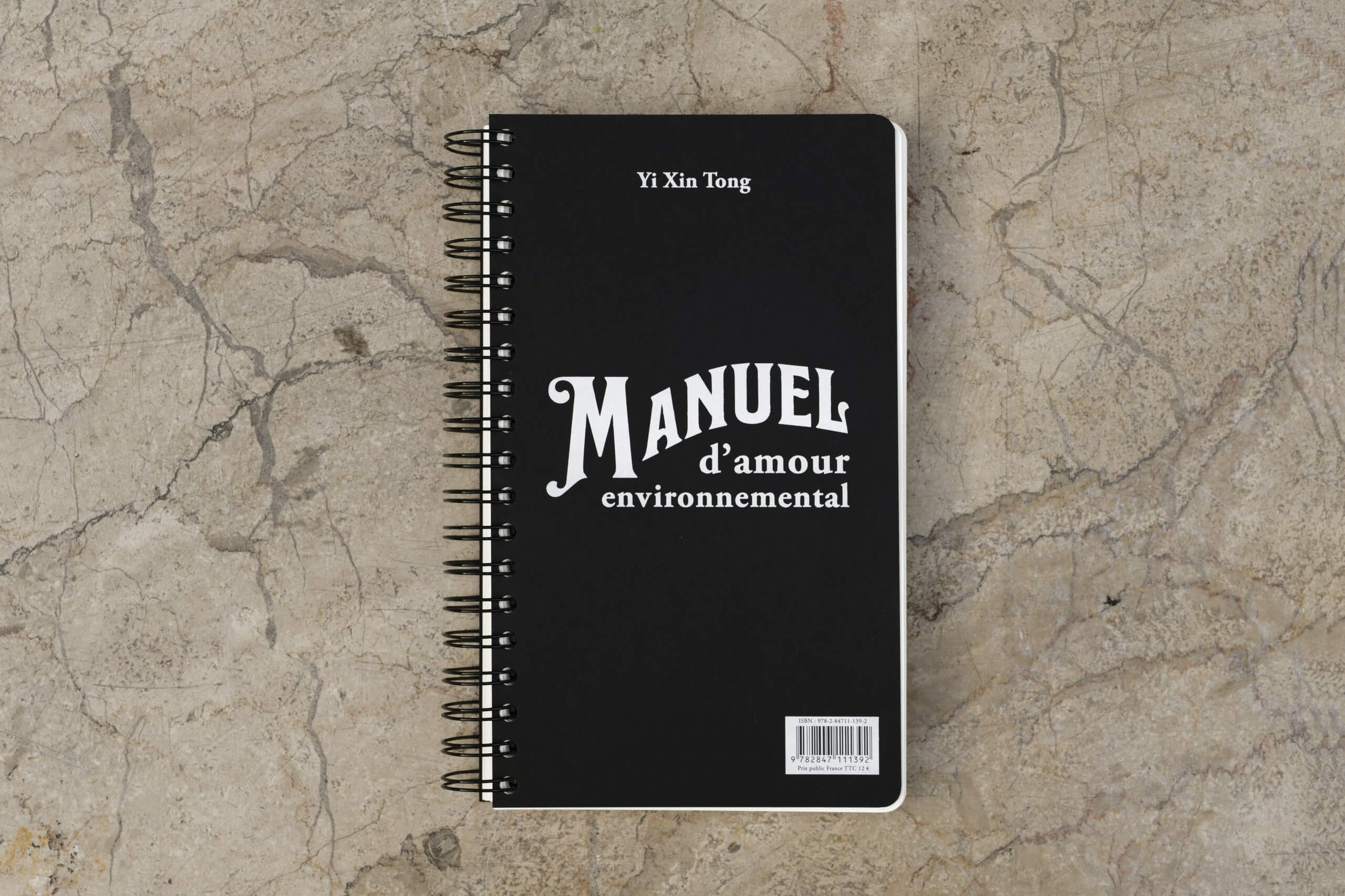 Un cahier à spirale noir avec le titre Manuel d'amour environnemental et l'auteur Yi Xin Tong sur la couverture, posé sur une surface en pierre texturée et craquelée.