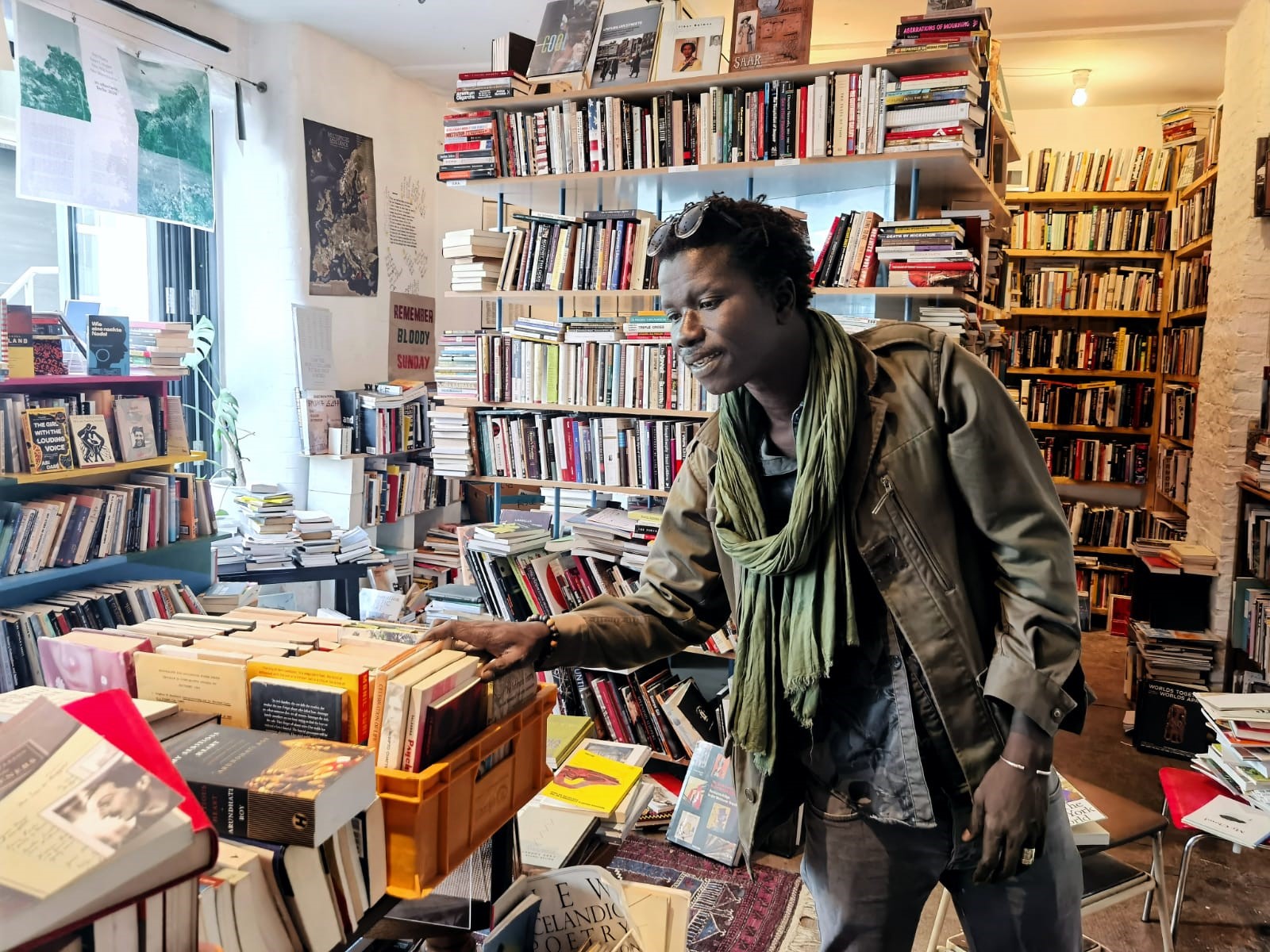 A person in a green scarf and jacket browses books in a crowded, well-lit bookstore with shelves full of books and stacks of books on tables. The atmosphere appears cozy and eclectic.