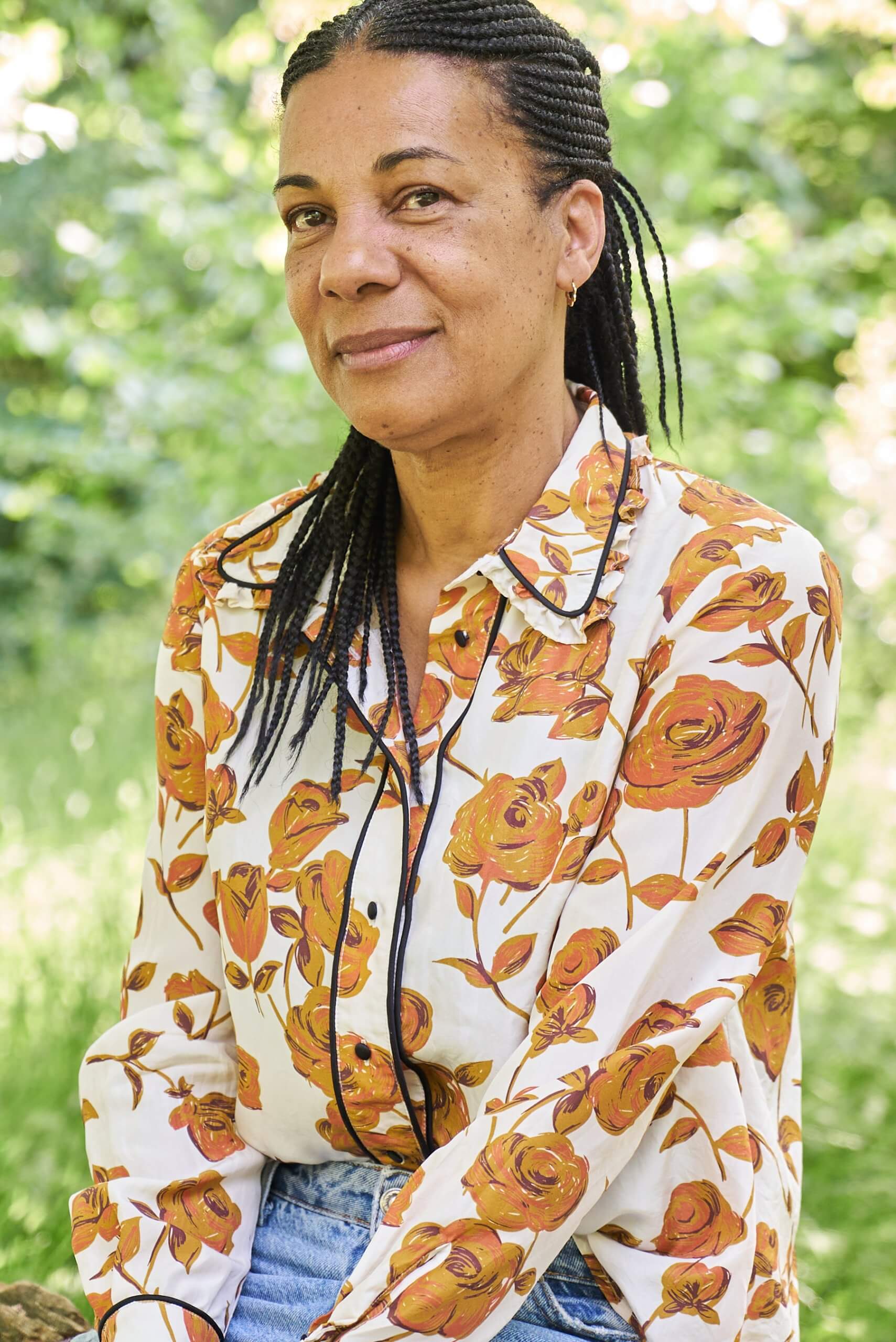 Une femme aux cheveux tressés est assise dehors, vêtue d'un chemisier blanc à motifs floraux orange et d'un jean bleu. Elle regarde l'objectif avec un léger sourire. Un feuillage vert est flou en arrière-plan.