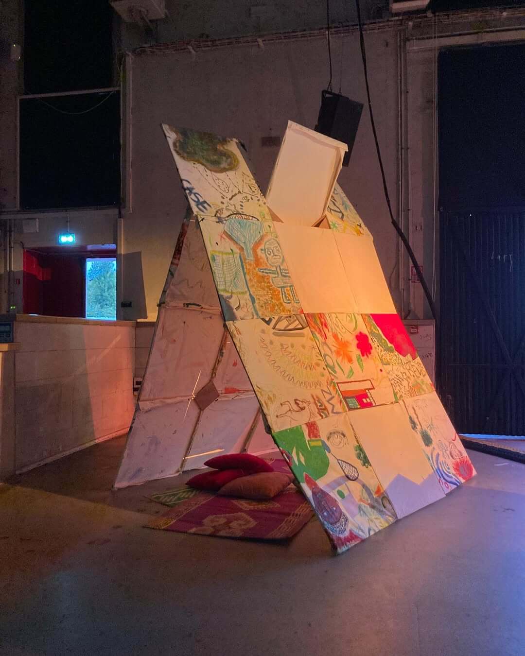 Une grande structure en forme de tente, composée de panneaux colorés recouverts de divers dessins et motifs, est installée à lintérieur. Deux coussins rouges sont placés sur un tapis devant elle. Larrière-plan représente un espace industriel avec une porte fermée.