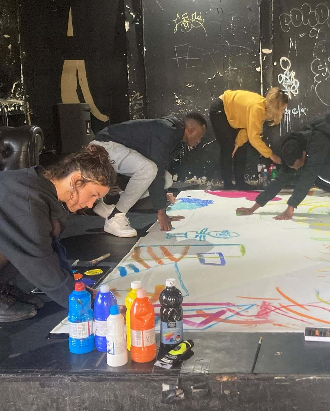 Quatre personnes sont penchées sur une grande toile sur une scène sombre et peignent avec différentes couleurs. Des bouteilles de peinture sont visibles au premier plan. À l'arrière-plan, un mur avec des graffitis.