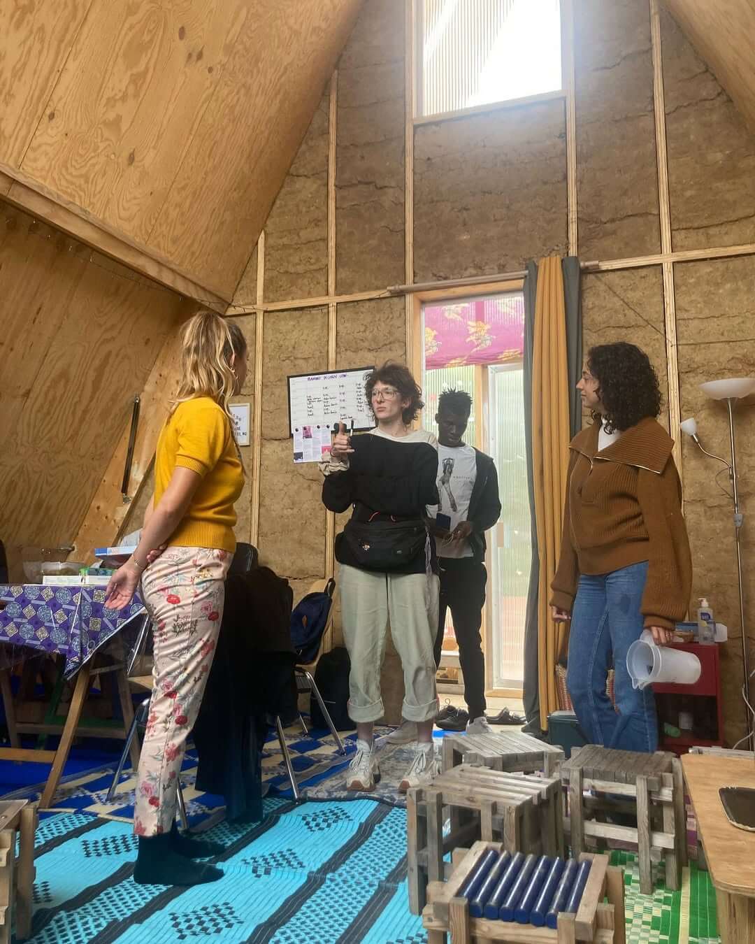 Quatre personnes se tiennent debout dans une cabane en bois en forme de A dont lintérieur est inachevé. Une personne tient une caméra, tandis que dautres écoutent. Une table avec une nappe colorée et un tableau blanc est visible. La lumière entre par une grande fenêtre à larrière-plan.