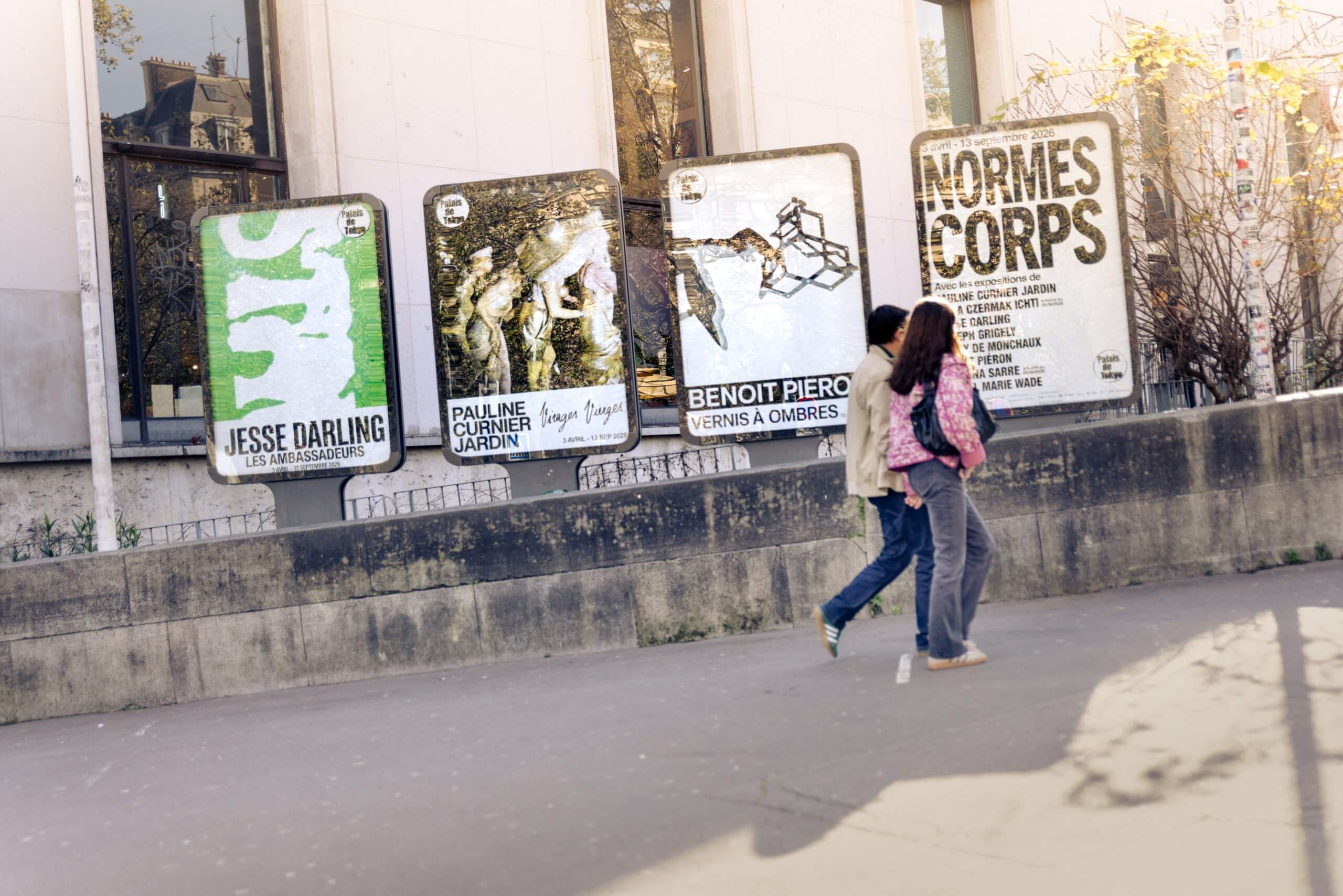 Deux personnes passent devant des affiches d'expositions artistiques placées sur un mur en béton dans un environnement urbain.