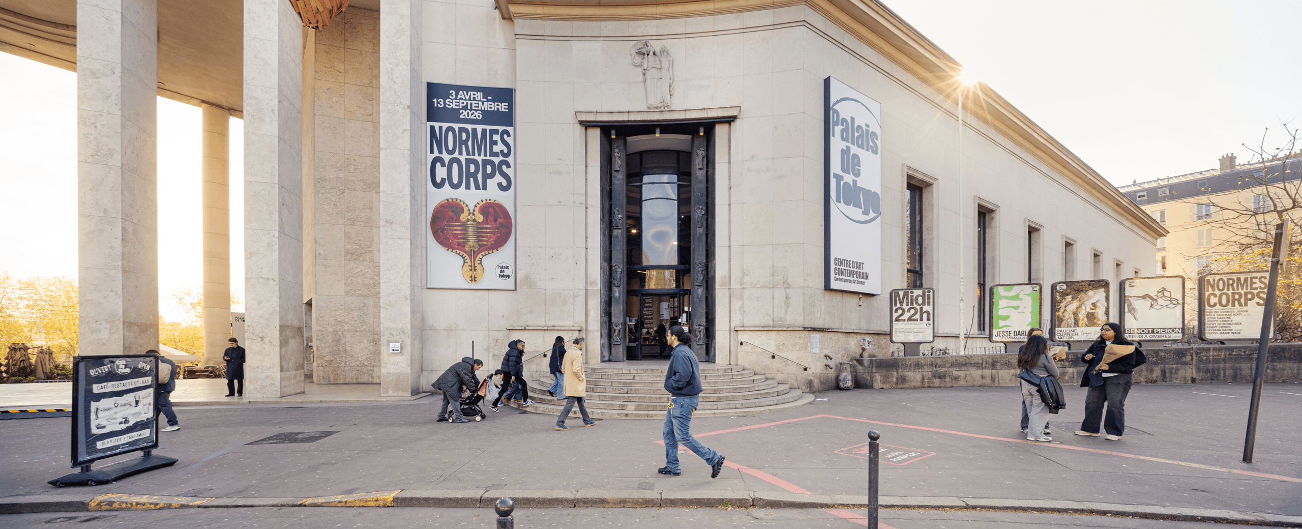 Des personnes marchent à l'extérieur d'un grand bâtiment de musée avec des bannières annonçant l'exposition Normes du Corps, datée du 3 avril au 13 septembre 2023.