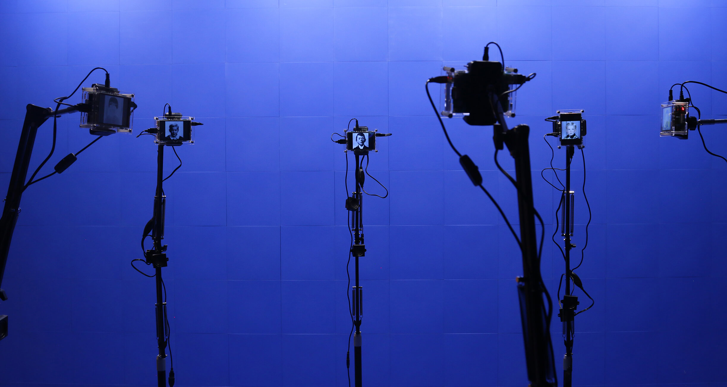 Plusieurs appareils montés sur des trépieds dans une pièce à fond bleu, chacun affichant une image en noir et blanc d'un visage.