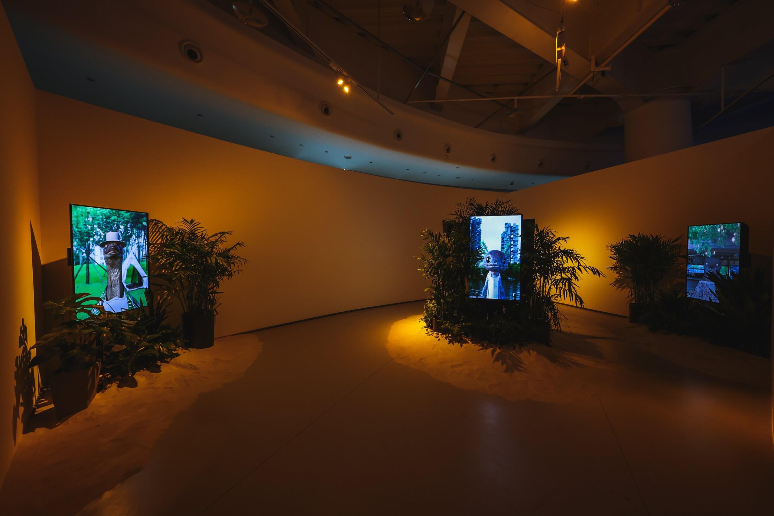 Installation vidéo dans une galerie avec des écrans entourés de plantes, projetant des images de personnages stylisés dans un éclairage doux et chaud.