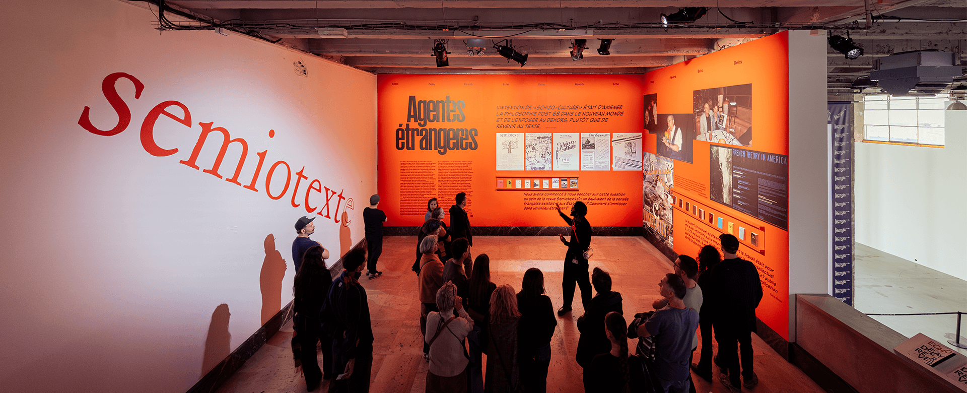 Un groupe de personnes visite une exposition d'art dont les grands murs rouges affichent les mots semiotext(e) et Agents étrangers, ainsi que divers textes et images.