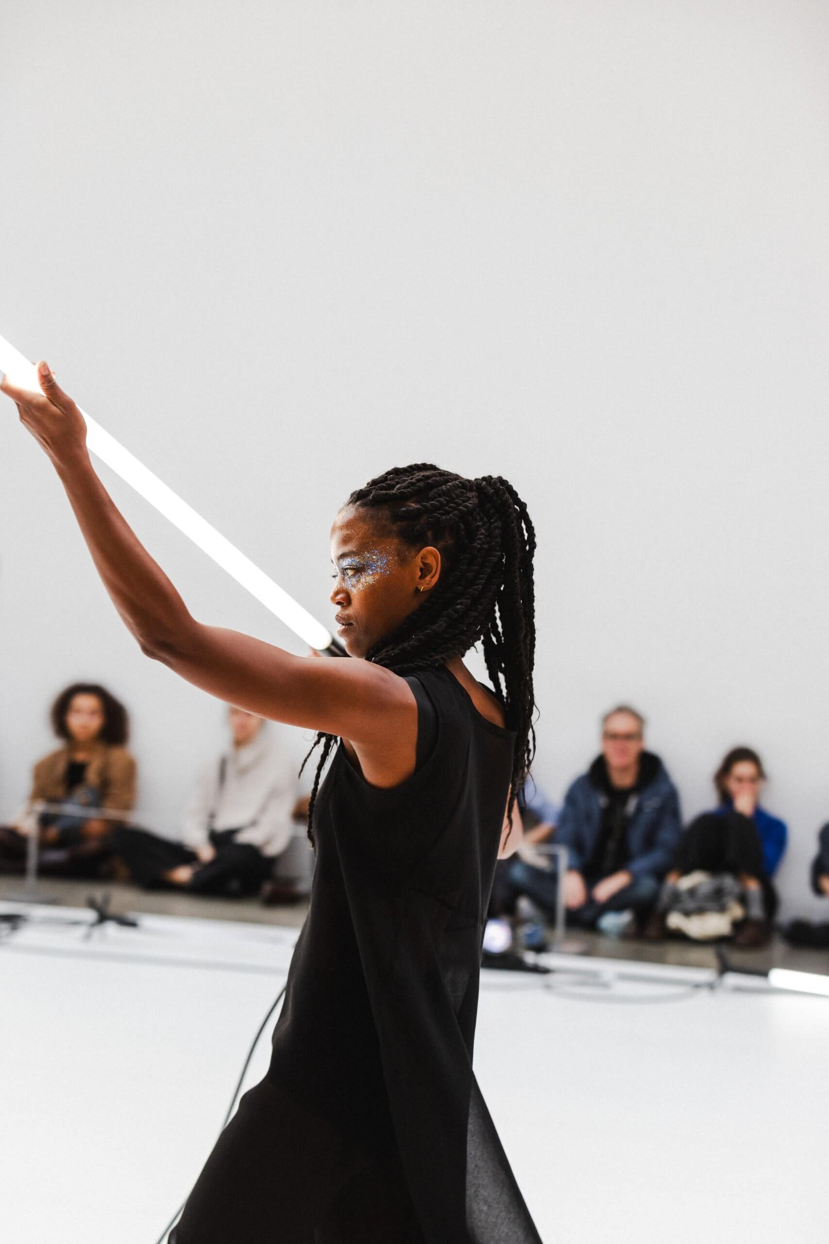 Une femme en robe noire tient un long bâton, se tenant droite avec une expression confiante.