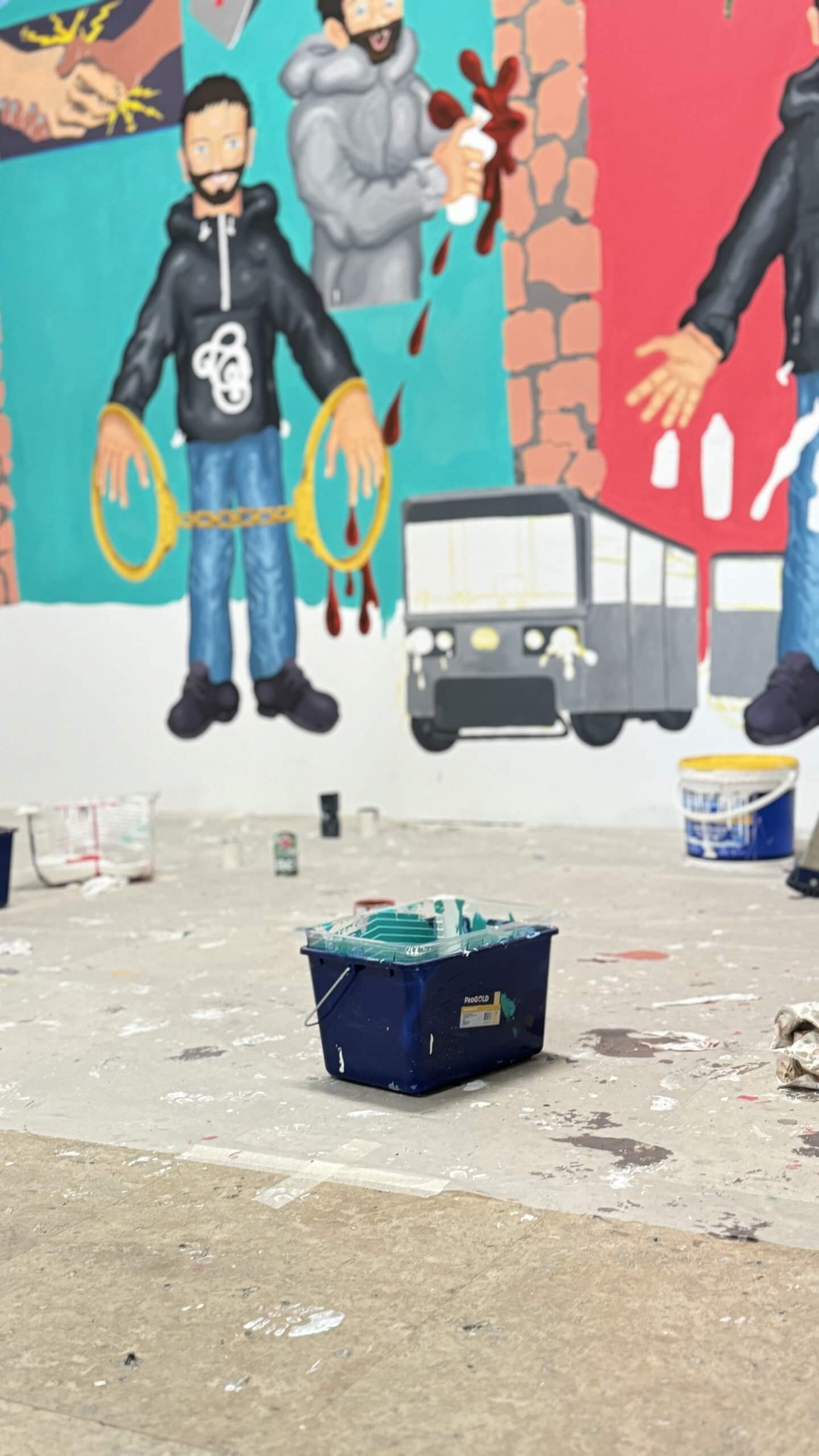 Blue paint bucket on the floor in front of a mural of characters and a bus.