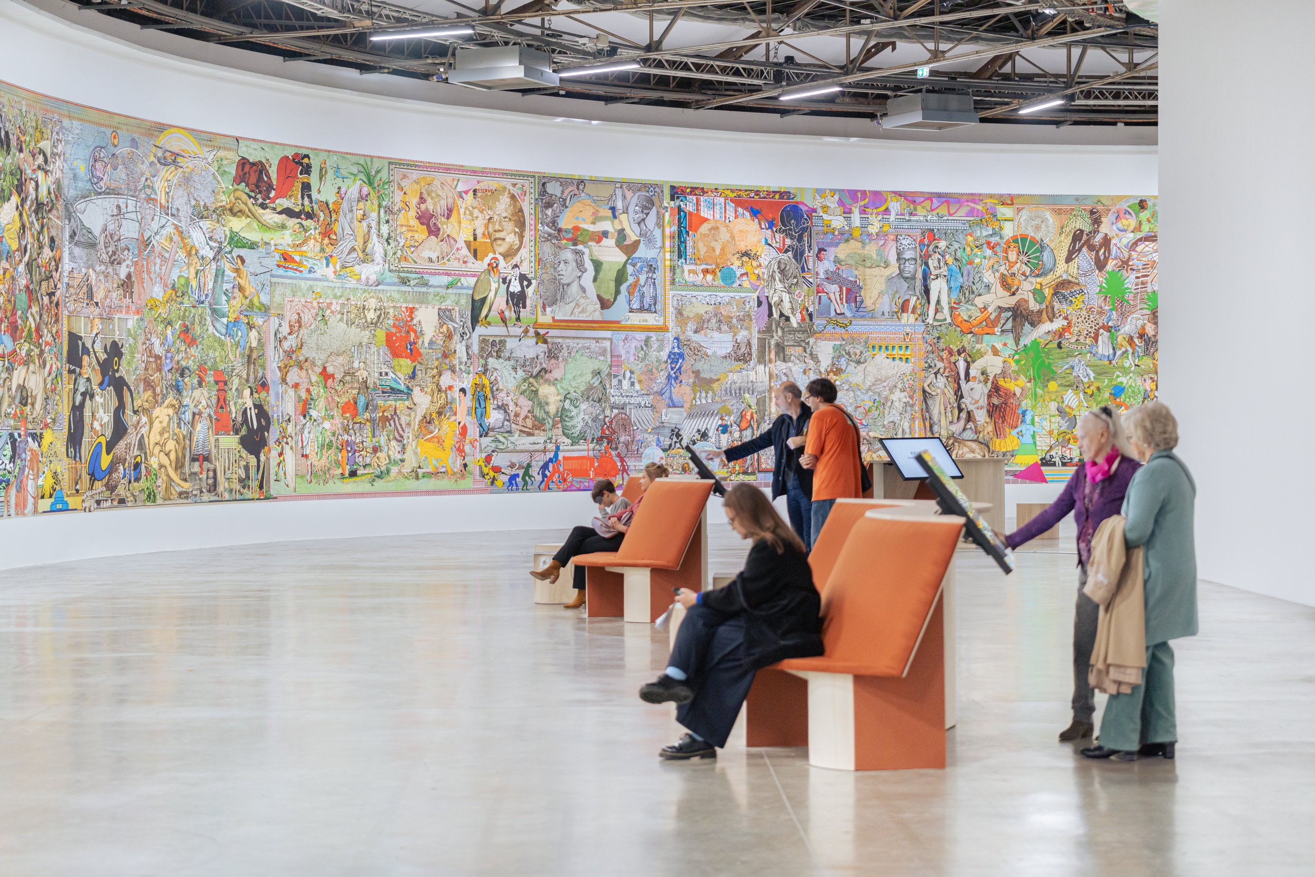 Les visiteurs admirent une grande œuvre colorée dans une galerie dart. Lœuvre, de la taille dun mur, est remplie de scènes complexes et détaillées. Plusieurs visiteurs sont assis sur des bancs, tandis que dautres sont debout, interagissant avec des écrans daffichage. Lespace est ouvert et bien éclairé.