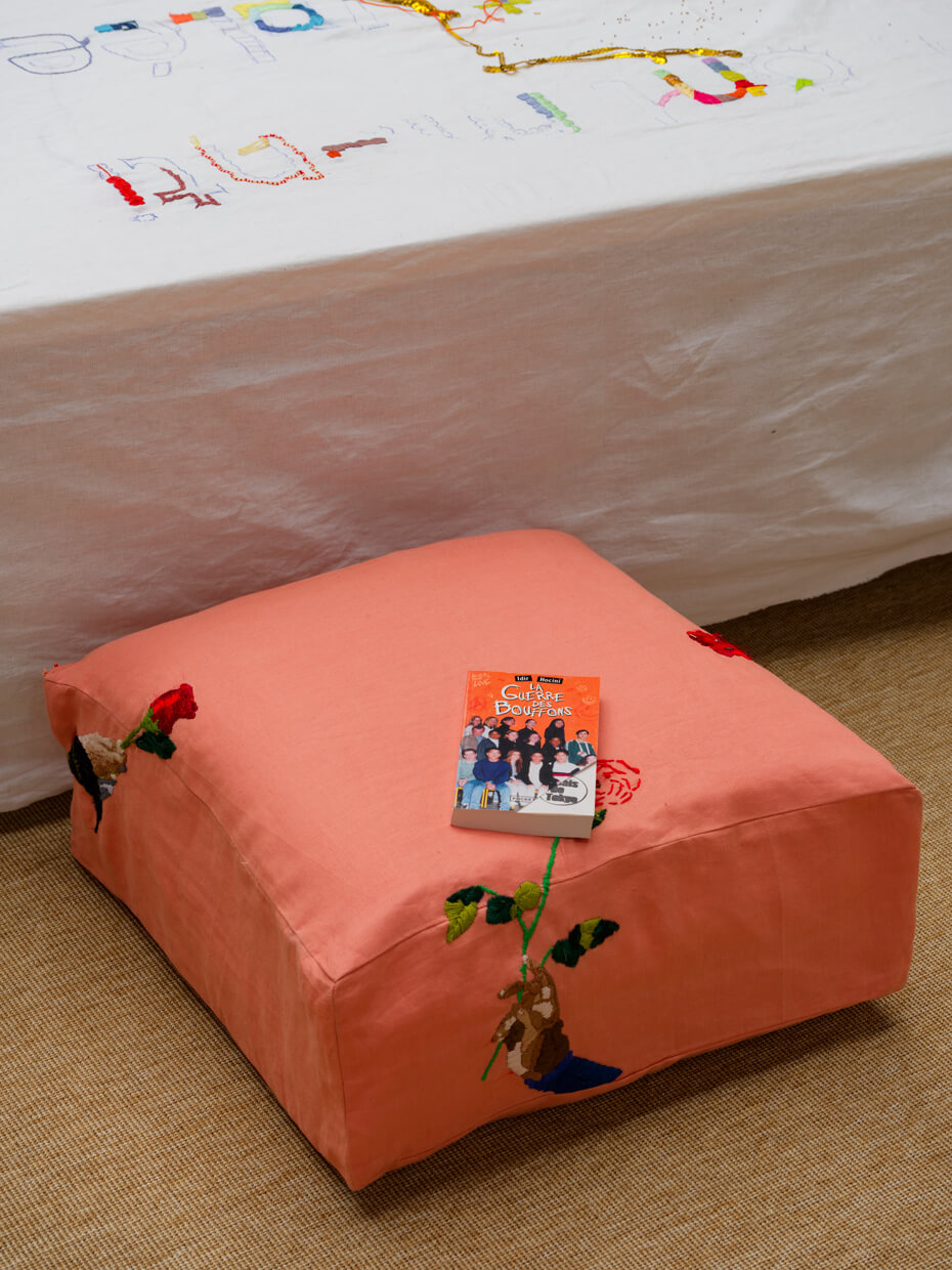 Un coussin carré rose avec broderie florale repose sur un tapis beige. Sur le dessus du coussin se trouve un livre avec une couverture orange vif représentant un groupe de personnes. Derrière le coussin se trouve un tissu blanc avec des motifs colorés.