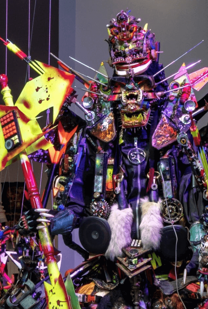 Une sculpture complexe et colorée présente une figure humanoïde ornée de divers éléments métalliques, électroniques et en tissu. La figure porte un masque tribal, des couleurs vives et tient des objets abstraits, créant une apparence complexe et éclectique.