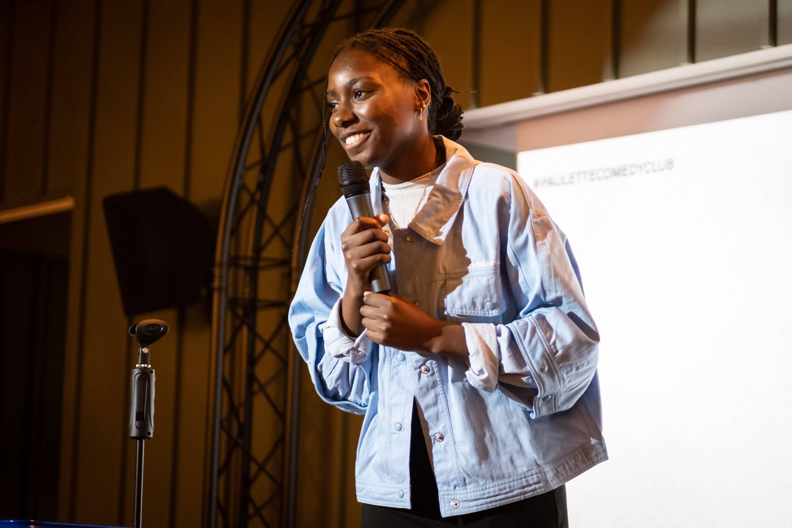Une personne se tient debout sur scène, tenant un micro, vêtue d'une veste bleu clair sur une chemise blanche. Elle sourit et semble parler. Un écran de projection est partiellement visible en arrière-plan.