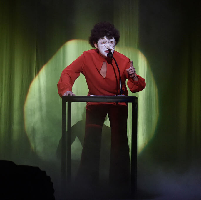 Une personne au visage peint en blanc et en tenue rouge se tient derrière un podium et parle dans un microphone. Des rideaux verts et un éclairage ambiant créent une atmosphère dramatique. La personne fait un léger geste d'une main.