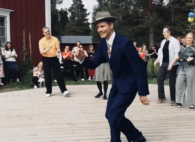 Une personne en costume bleu et chapeau gris danse avec énergie sur une terrasse en bois. Une foule de gens regarde en arrière-plan, certains souriant et applaudissant, près d'un bâtiment rouge. Des ballons sont visibles à droite.