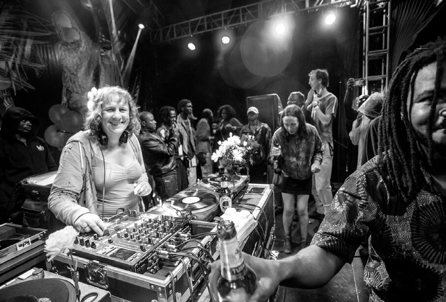 Une photo en noir et blanc d'une DJ souriante qui utilise une table de mixage avec des platines lors d'un événement animé. Une foule est rassemblée derrière elle sur scène, profitant de la musique. Un homme tient une bouteille au premier plan. Les lumières de la scène illuminent la scène.