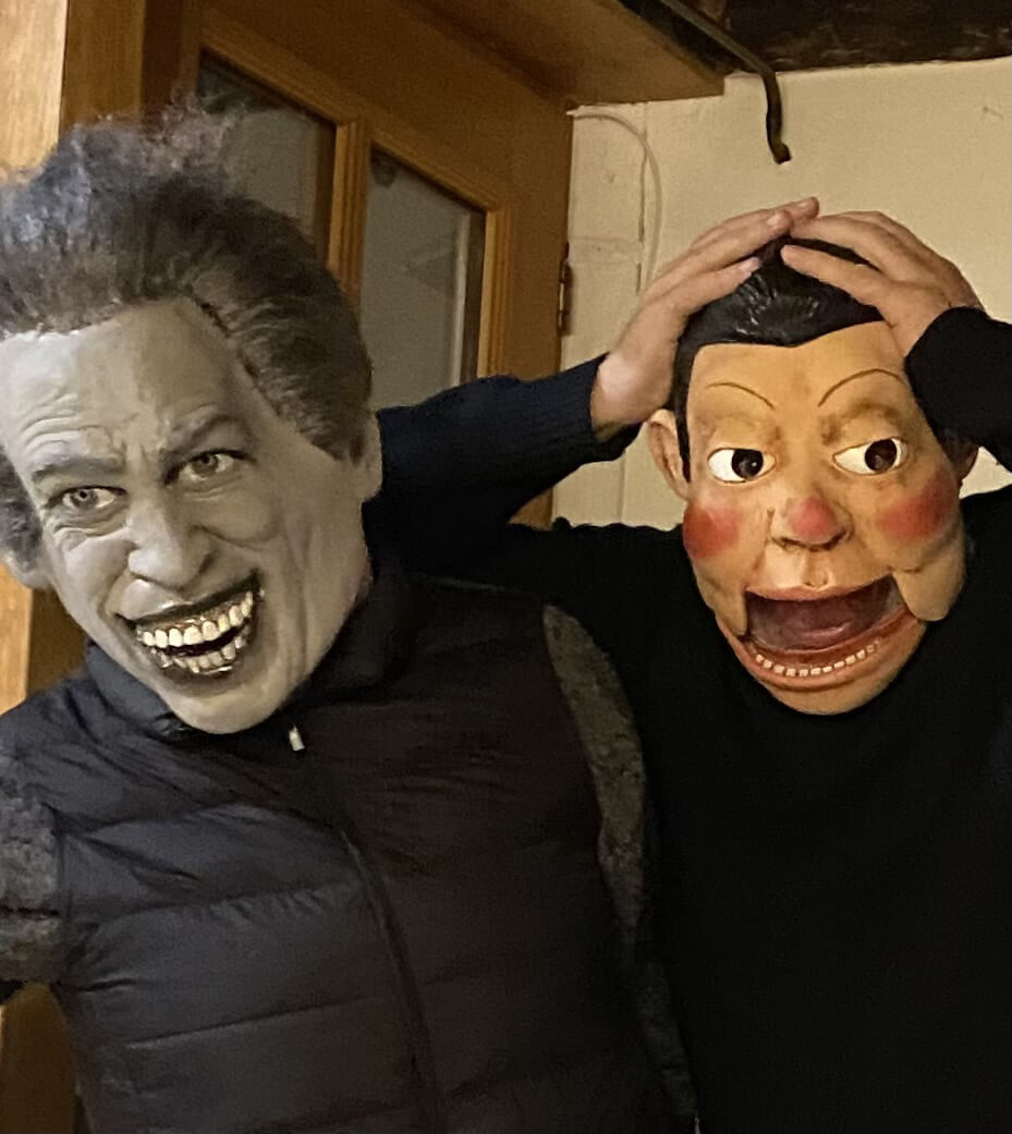 Deux personnes portant des masques posent à l'intérieur. La personne de gauche porte un masque avec des traits exagérés et un large sourire. La personne de droite porte un masque avec la bouche ouverte et les joues roses, tenant sa tête à deux mains.