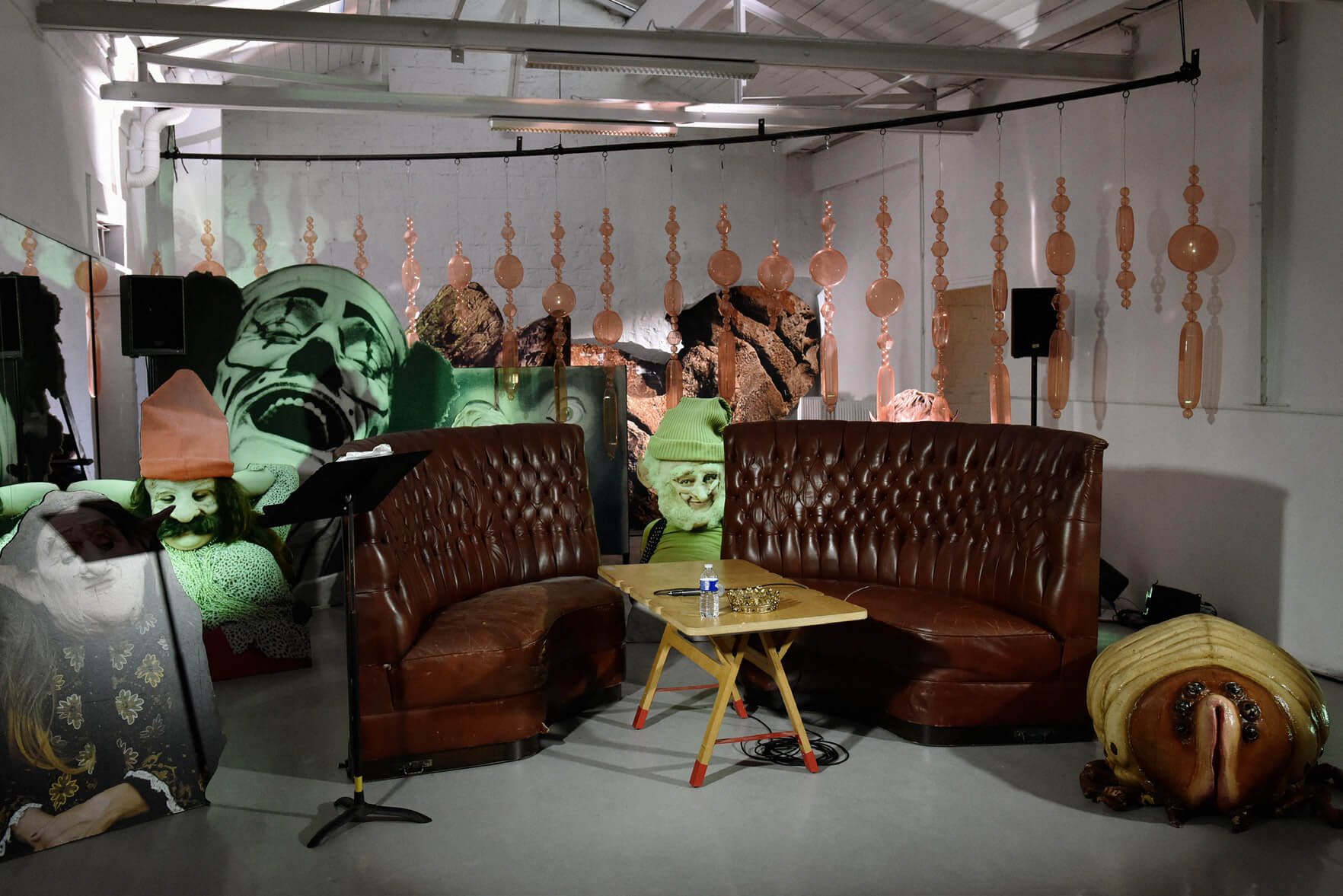 Une pièce au décor surréaliste comprend deux canapés en cuir marron, une petite table en bois et diverses grandes images de visages et de formes abstraites. Des objets suspendus et des murs à motifs créent une atmosphère éclectique.