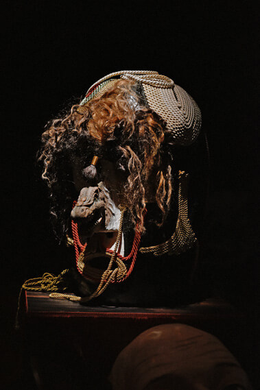 Une œuvre sculpturale composée d'un mélange de matériaux, notamment des cordons tressés, une tête texturée ressemblant à un animal et des éléments ressemblant à des cheveux emmêlés, est exposée sur un piédestal sur un fond sombre.