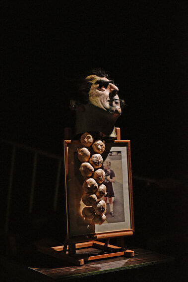 Scène faiblement éclairée représentant un buste posé sur un socle en bois. Sous le buste se trouvent plusieurs objets ronds ressemblant à des bulbes d'ail. Au-dessous, une photographie encadrée représente une personne vêtue de noir, debout sur une plage, avec de l'eau en arrière-plan.
