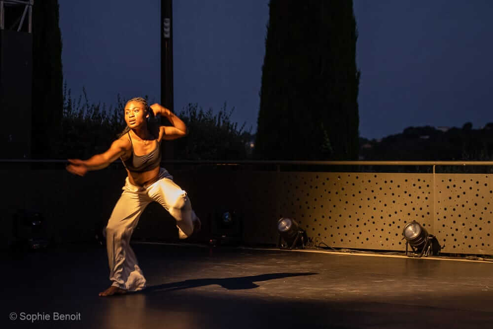Une personne danse avec énergie sur une scène faiblement éclairée la nuit. Le danseur porte un haut sombre et un pantalon clair. Les lumières de la scène sont positionnées sur le sol et l'arrière-plan est sombre avec des silhouettes d'arbres.