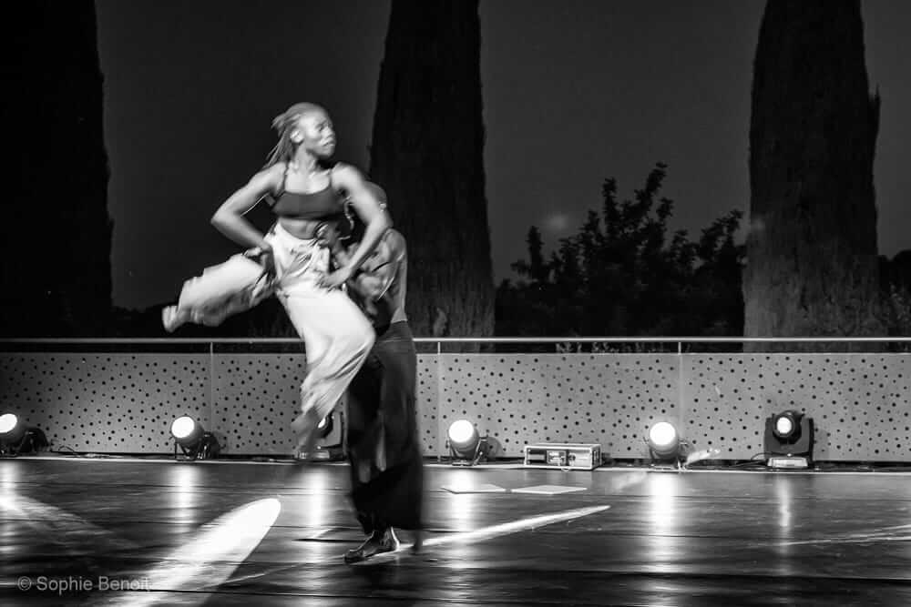 Sur cette photographie en noir et blanc, deux danseurs se produisent sur scène la nuit. L'un d'eux est soulevé dans les airs par son partenaire. Des arbres et un éclairage de scène sont visibles en arrière-plan.