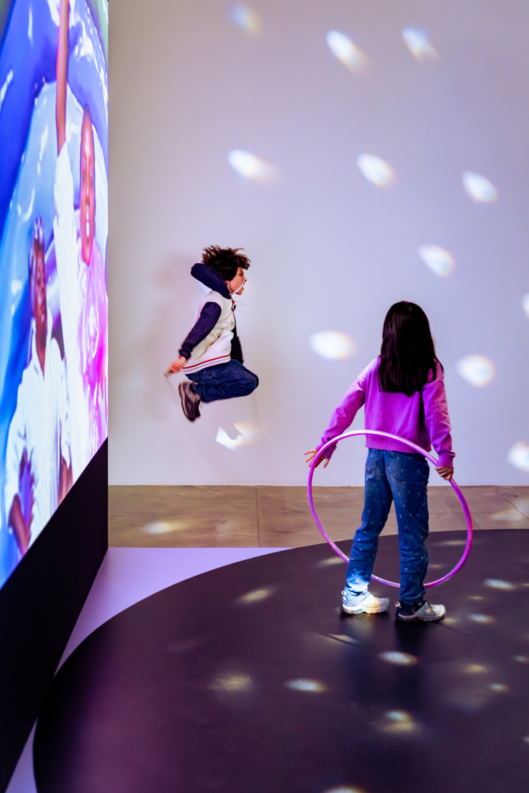 Deux enfants jouent dans une pièce éclairée par des projecteurs lumineux colorés. L'un saute près d'un écran affichant des personnages, tandis que l'autre tient un cerceau violet. Le sol est sombre et parsemé de points lumineux.