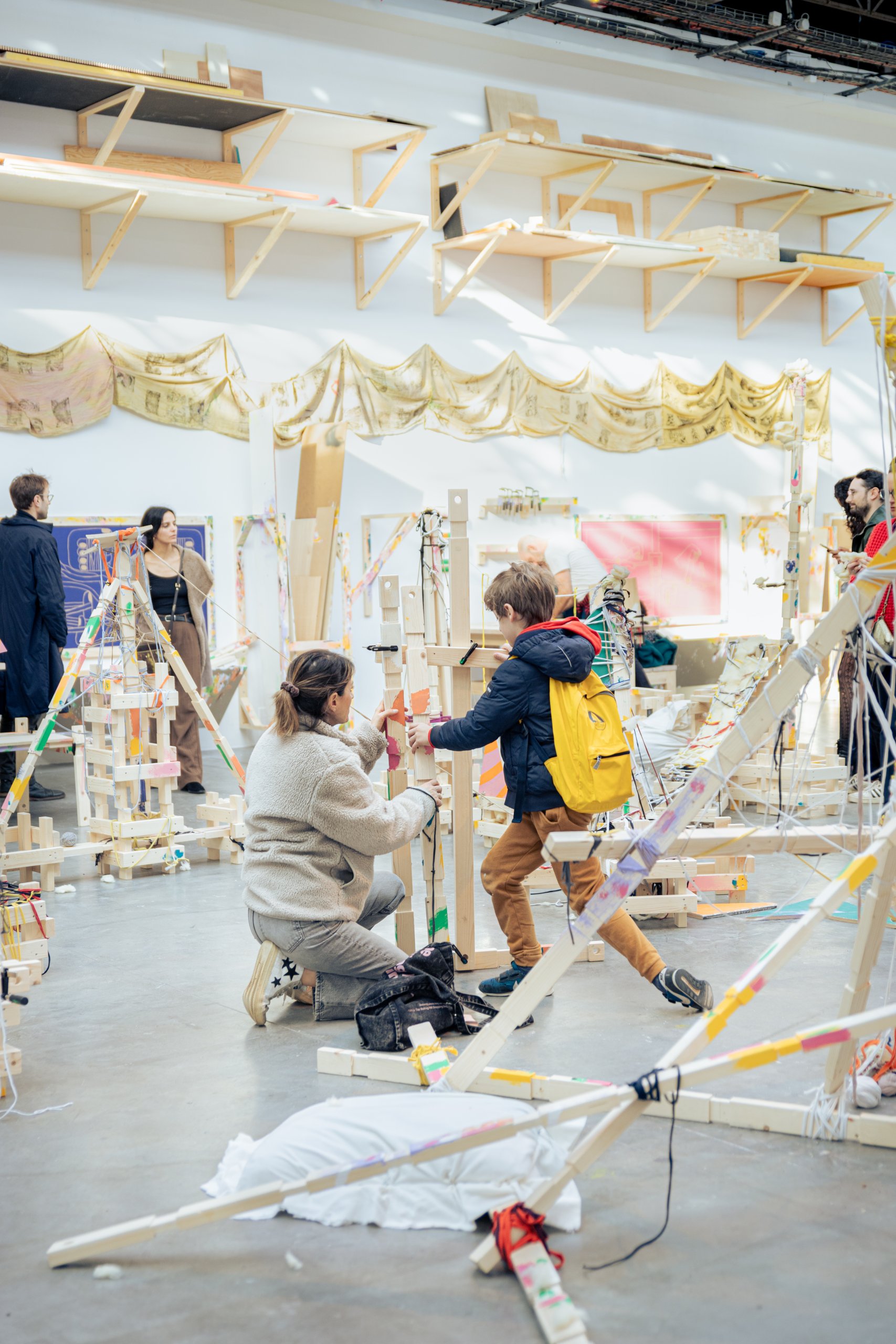 Dans une salle spacieuse, des spectateurs construisent et interagissent avec une grande installation artistique abstraite. L'installation présente des structures en bois ornées de matériaux colorés. Plusieurs personnes participent au processus créatif et explorent l'œuvre.