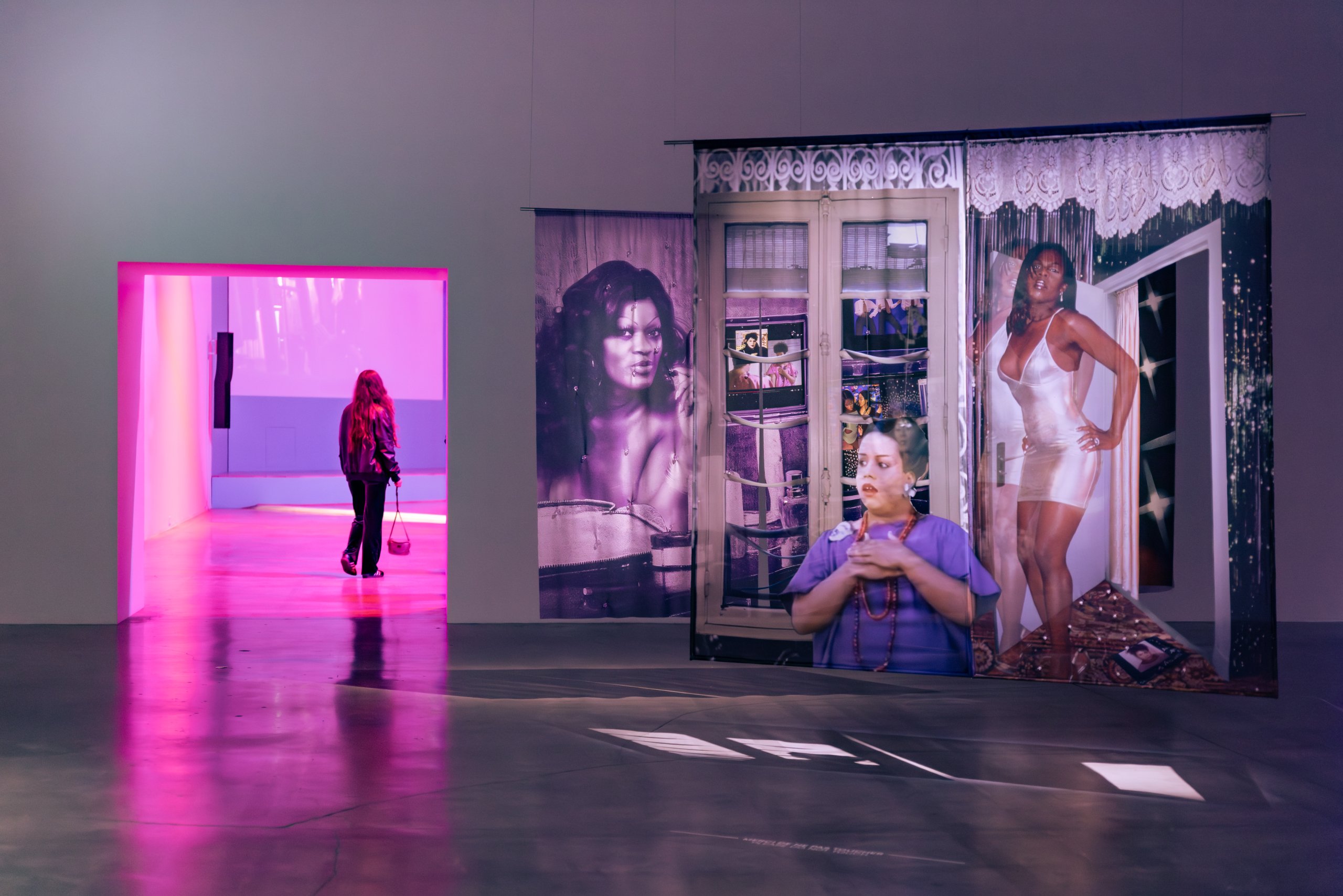 Une personne se tient dans une galerie d'art, partiellement derrière de grands panneaux photographiques représentant des femmes en tenue. La pièce est éclairée de rose par une lumière provenant d'une zone adjacente où une autre personne est visible. Le sol est réfléchissant.