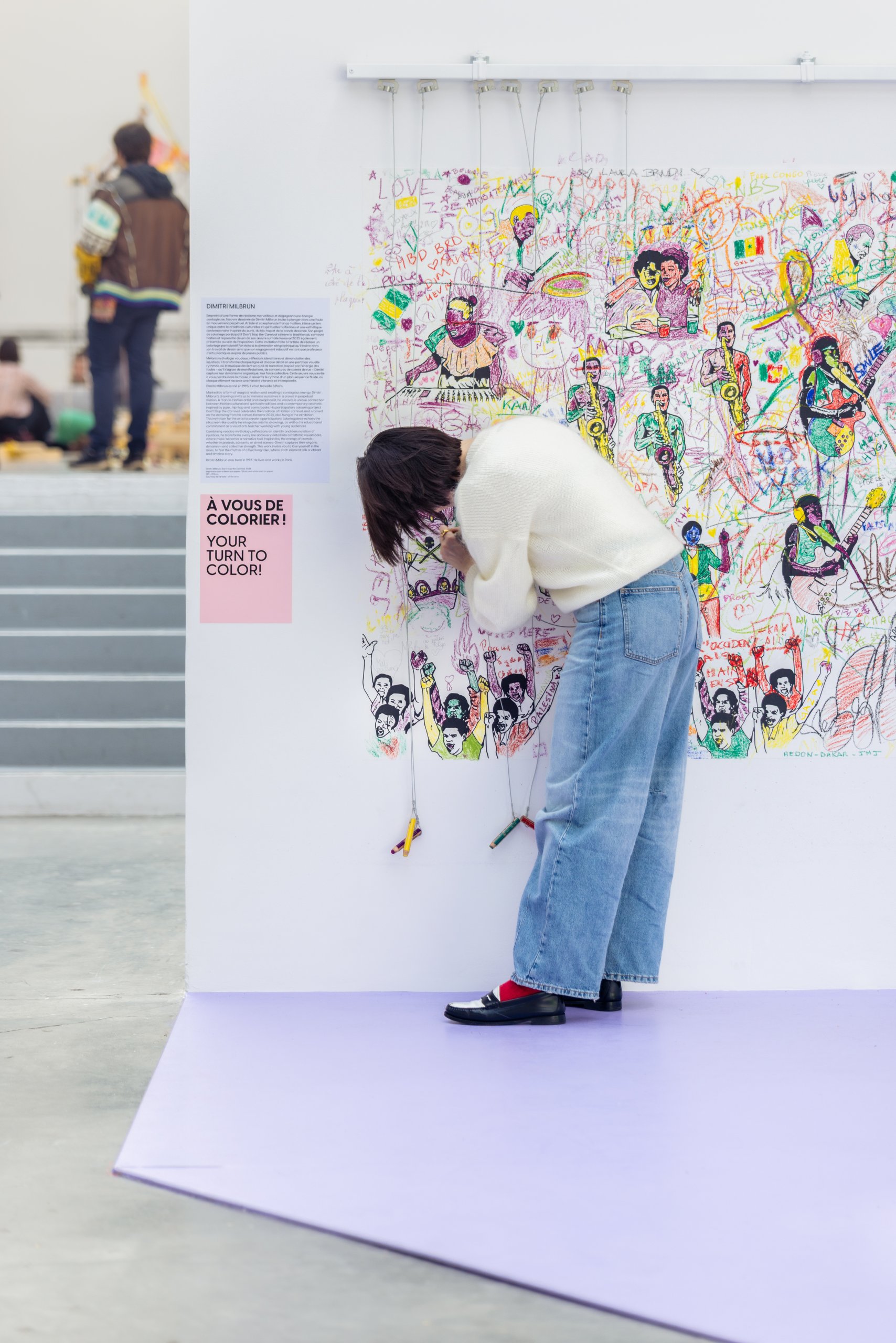 Une personne se tient devant un écran mural interactif coloré, ajustant ou examinant l'œuvre. Elle porte un pull blanc, un jean et des chaussures noires avec des chaussettes rouges. Le mur est orné de dessins et de textes. Le décor évoque une galerie ou un espace public.
