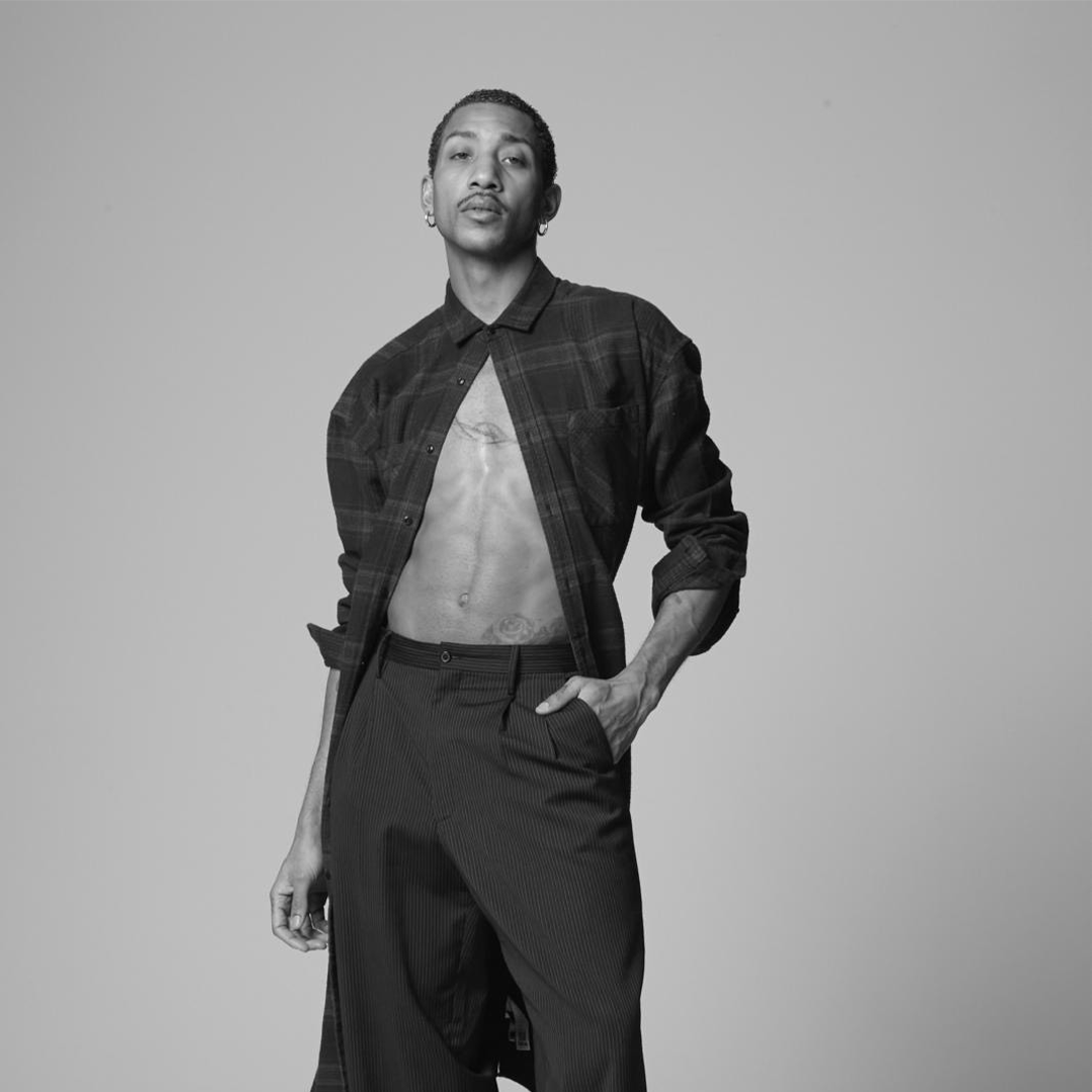 Une personne se tient debout sur un fond uni, vêtue d'une chemise à carreaux ouverte qui dévoile sa poitrine et d'un pantalon à rayures fines taille haute. Une main est dans la poche de son pantalon, l'autre à ses côtés. La photo est en noir et blanc.