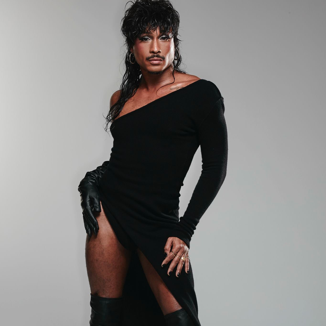 Une personne à la peau moyenne et aux cheveux foncés coiffés pose dans une robe noire à manches longues, à épaules dénudées et à fente haute, portant des gants noirs et des bottes hautes sur un fond gris uni.