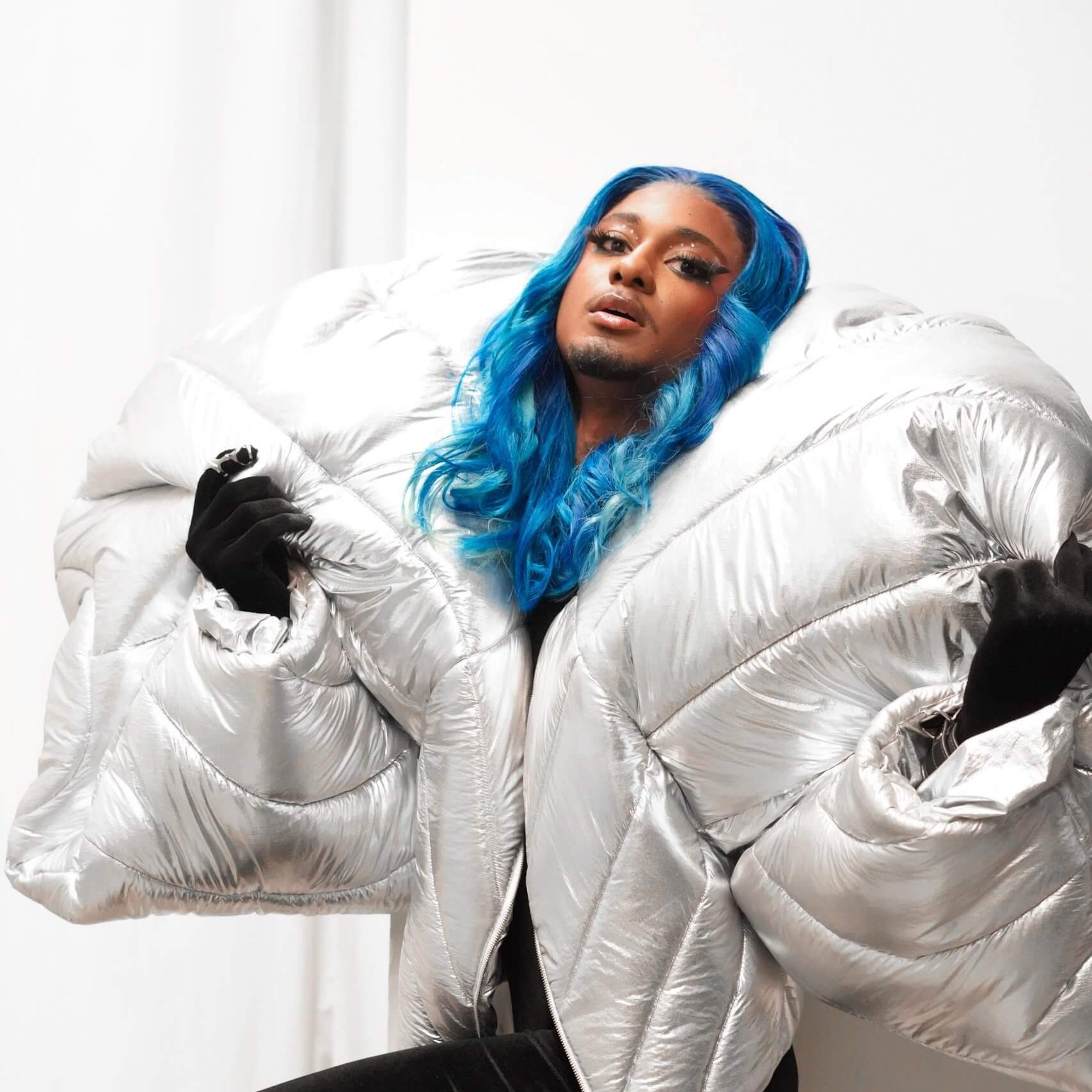 Une personne aux longs cheveux bleus porte une grande veste polaire argentée, des gants noirs et des bottes noires brillantes. Elle se tient debout sur un fond blanc.