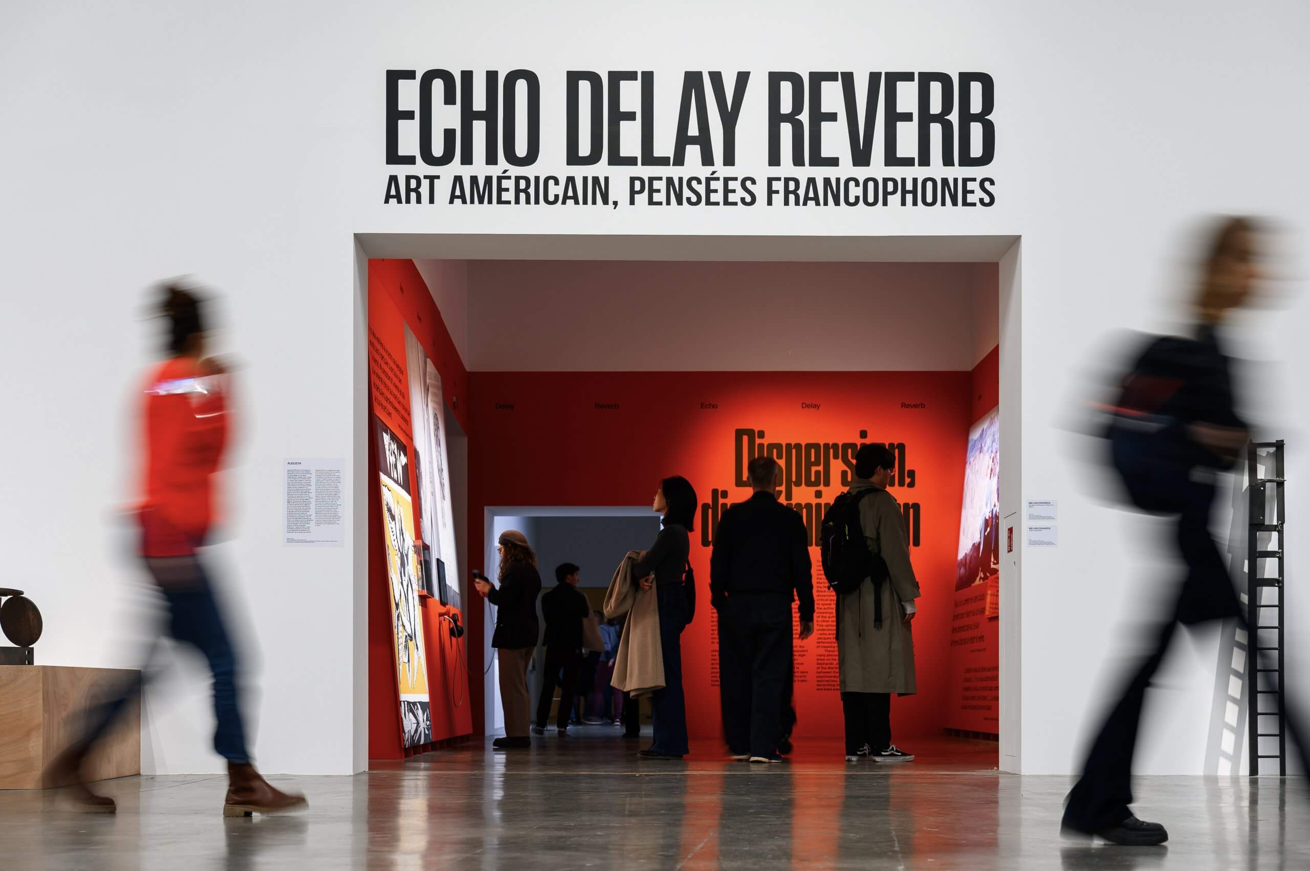 People stand and walk near the entrance of an art exhibit titled Echo Delay Reverb: Art Américain, Pensées Francophones, displayed on a white wall above a red-lit gallery space.