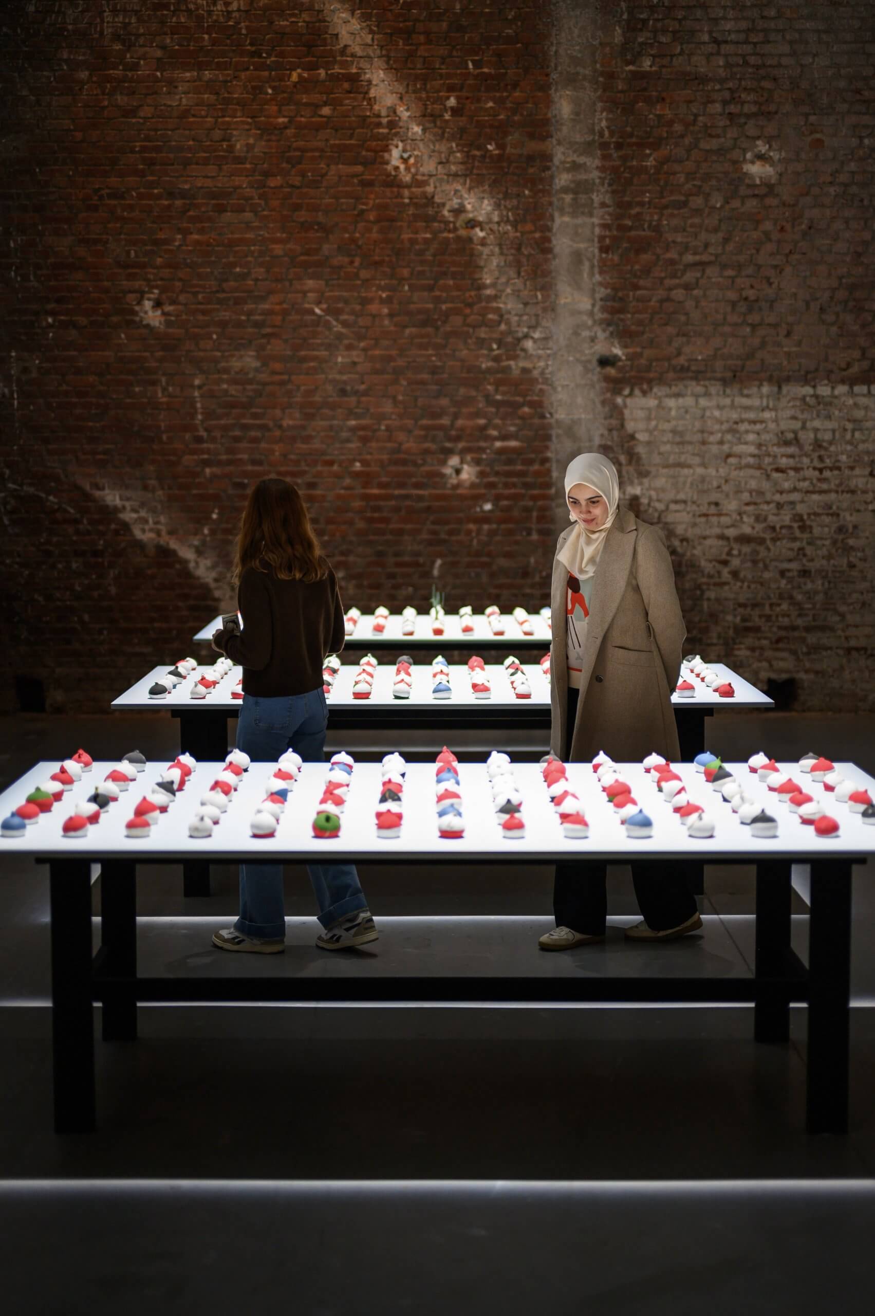 Two people observe rows of small colorful objects displayed on illuminated tables in front of a brick wall in an indoor setting.
