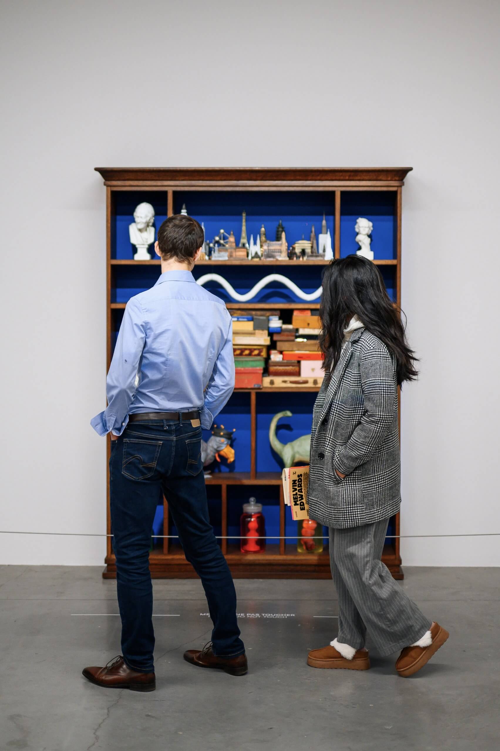 Two people stand in front of a wooden display shelf filled with various small statues, models, and objects against a blue background in a gallery setting.
