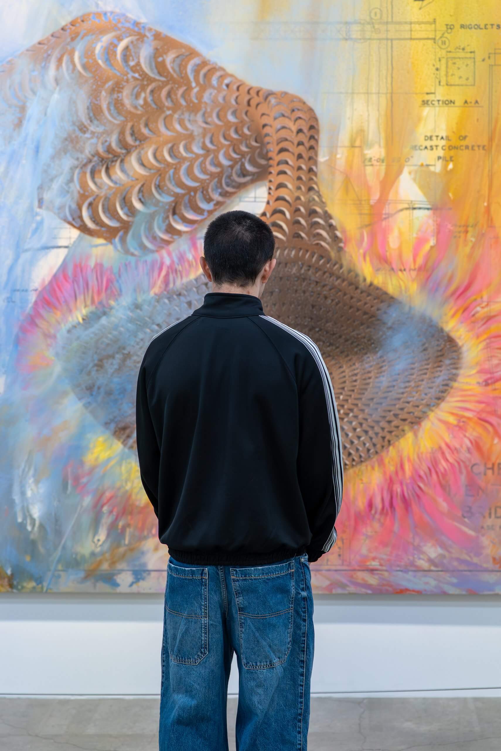 A person in a black jacket and blue jeans stands with their back to the camera, looking at a colorful abstract artwork on a gallery wall.