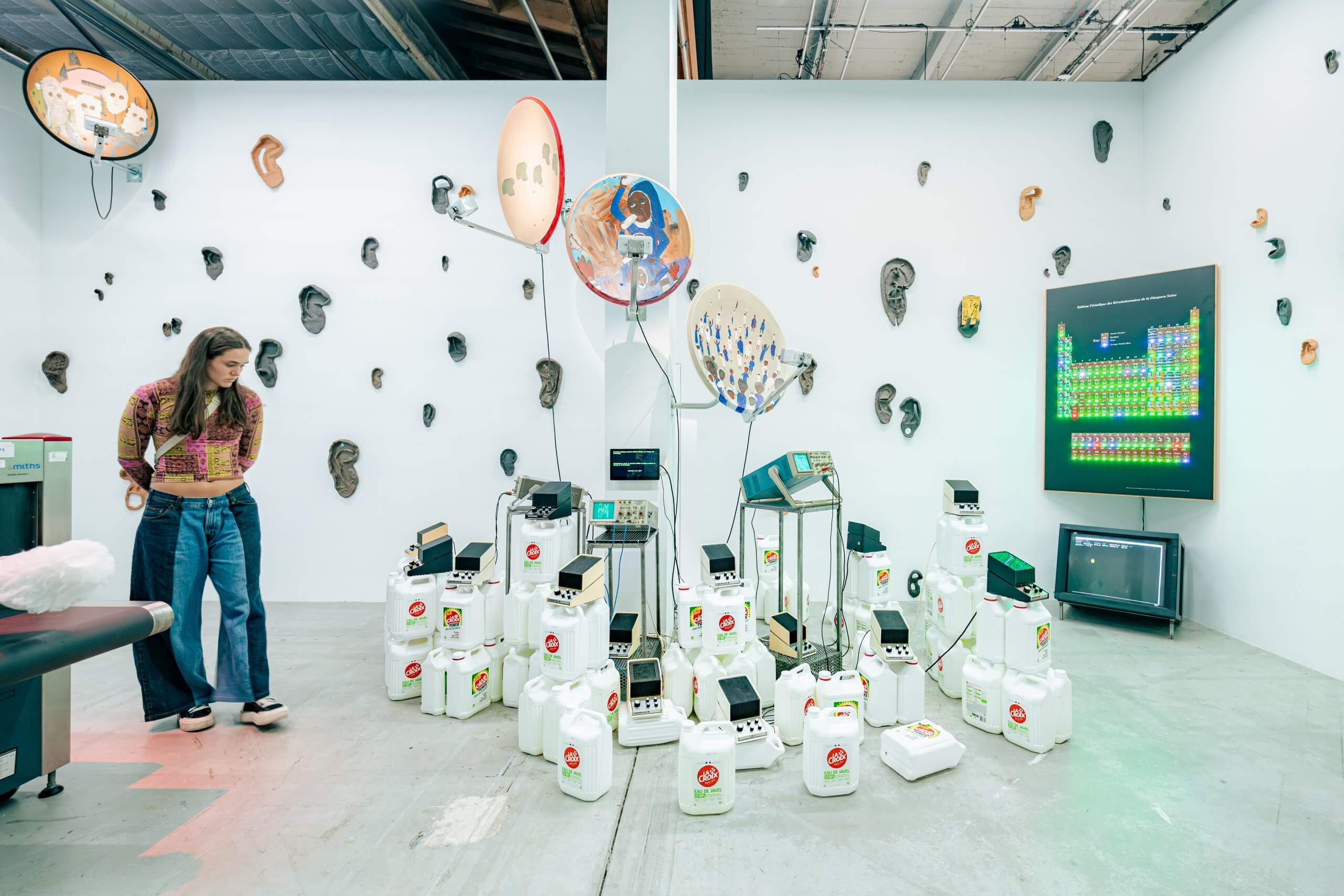 A woman stands in an art gallery with walls covered in ear sculptures and electronic devices arranged on white containers in the center of the room.