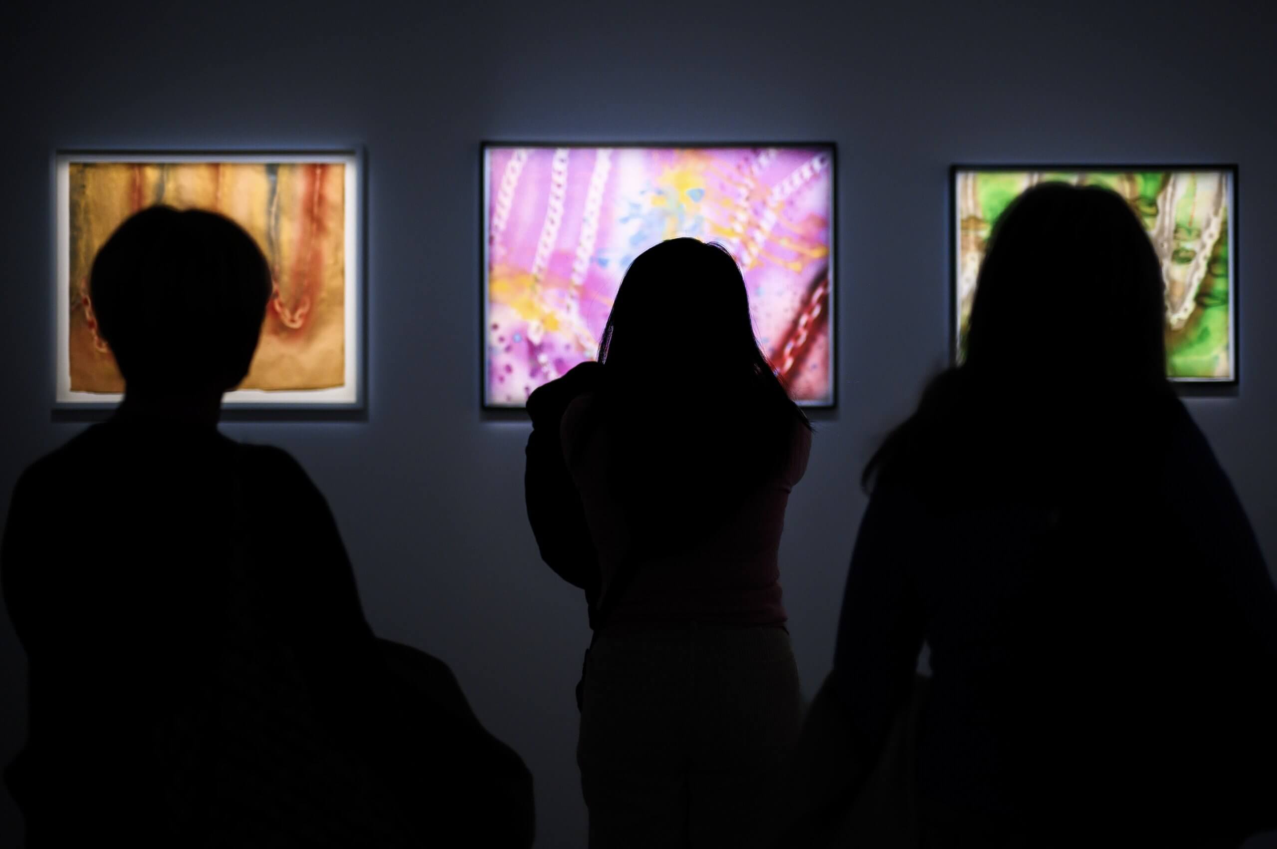 Silhouetted figures view three illuminated framed artworks on a dark wall in an art gallery.